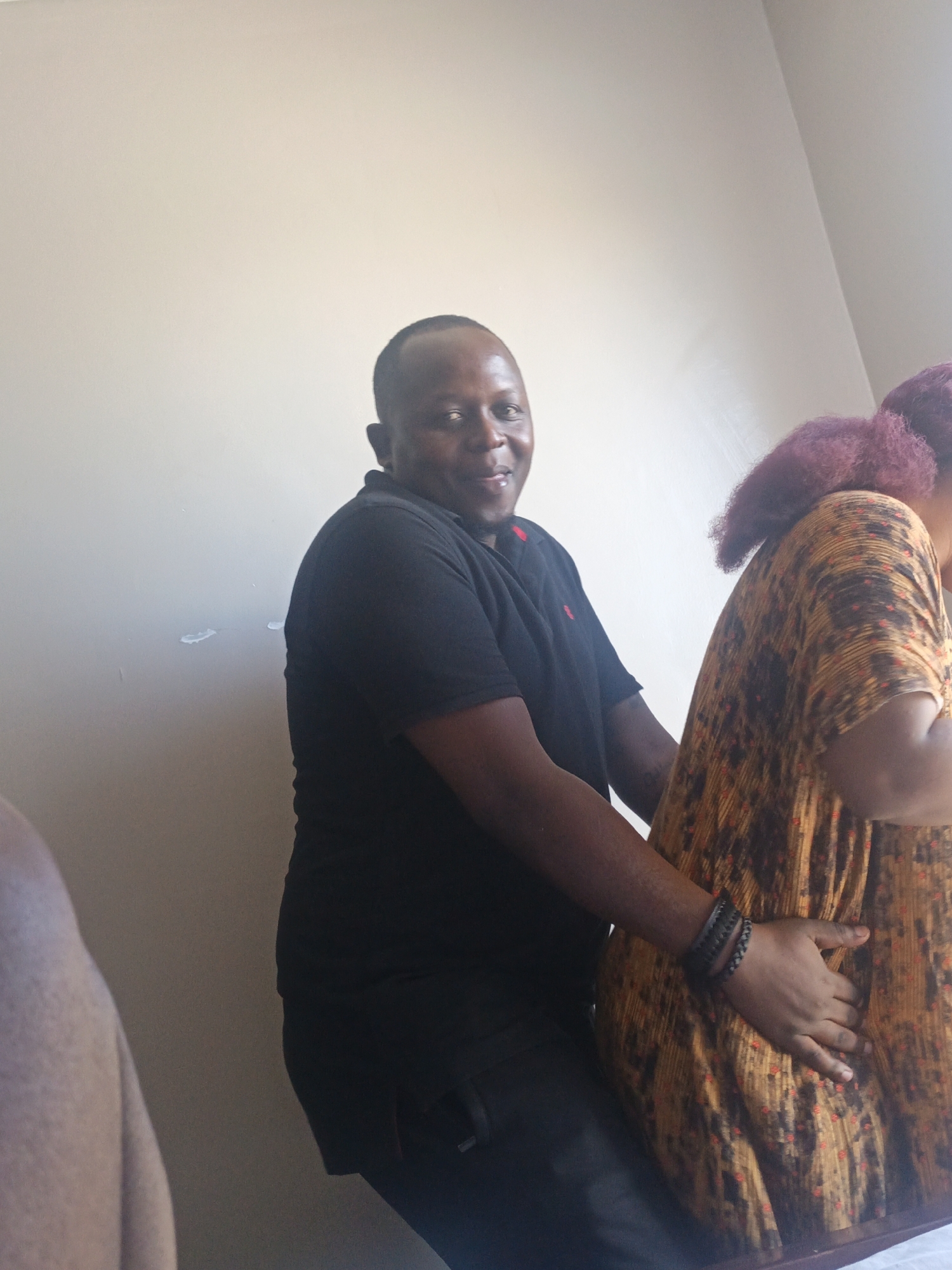 This image captures a candid indoor moment featuring two individuals, a man and a woman, in what appears to be Nairobi, Kenya. The man, positioned on the left, has short dark hair and is wearing a black polo shirt with a small red logo on the chest. He smiles warmly, looking directly at the camera. His left hand rests gently on the lower back of the woman beside him, and he wears a dark, possibly braided, bracelet on his wrist.
The woman is mostly seen from behind on the right side of the frame. Her hair is a distinctive reddish-purple color, styled in what appears to be twists or braids. She is wearing a patterned dress or top in hues of orange, brown, and black.
The background is a plain, light-colored wall, showing some minor scuff marks. The lighting is bright and even, suggesting it is daytime, possibly with natural light entering the room. A blurred arm or shoulder is visible on the far left, hinting at the presence of another person just out of frame. The overall impression is one of a casual, friendly interaction. No visible text is present in the image.