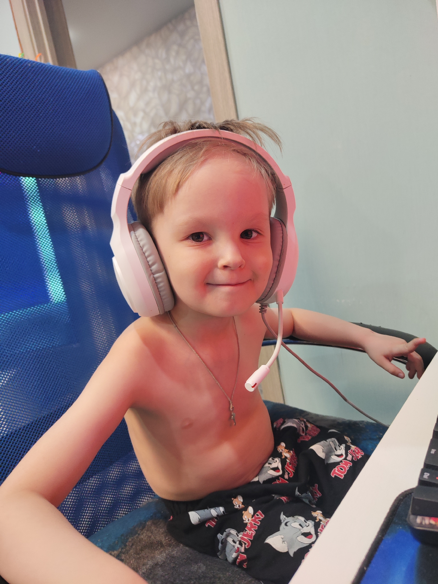 A young child, likely a boy, is seated in a blue mesh office chair, captured in a medium close-up indoors. He has light brown hair and is wearing pink and white over-ear headphones with an attached microphone, suggesting engagement in an online activity such as gaming, video calls, or virtual learning. The child is shirtless but wears black sleep shorts or pajamas adorned with various "Tom & Jerry" characters, with the text "TOM & JERRY" clearly visible on the fabric. A thin silver chain with a small pendant is visible around his neck.
The child looks directly at the camera with a gentle, pleasant smile, conveying a sense of happiness or contentment. His left arm rests on the chair's armrest, while his right hand is partially visible near a white desk surface and a black computer keyboard on the right side of the frame. The background features a simple, light blue-green wall and hints of light-colored wooden furniture or trim, indicating a domestic setting. The soft, even lighting suggests it is daytime. This scene of a child engaged with technology in a comfortable home environment is located in Chaikovsky, Russia.