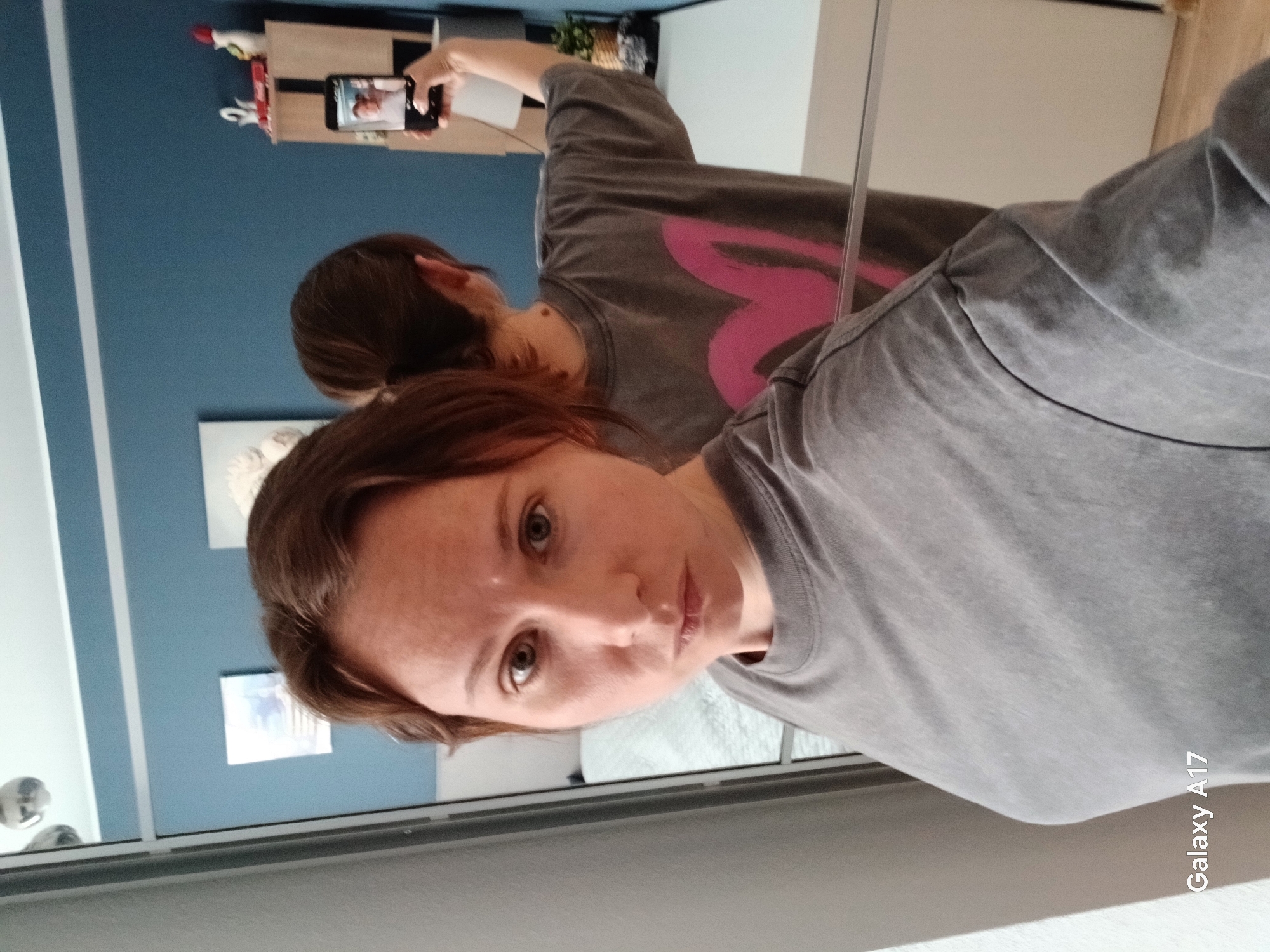A woman with short brown hair is taking a selfie in a mirror. She is wearing a gray t-shirt with a faded pink heart design. The reflection in the mirror shows her holding a smartphone up and taking the picture. The background includes a blue wall with framed artwork and a shelf with various items. The location is difficult to determine with the visual information provided, and there's no clear evidence to suggest this is Yekaterinburg, Russia.