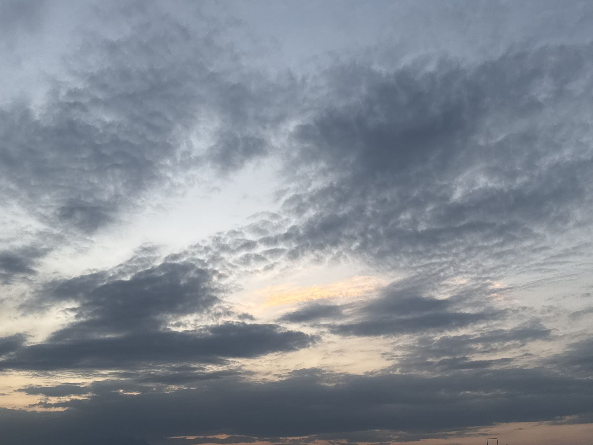 The image presents a broad view of an overcast sky, likely captured during either sunrise or sunset over San Francisco Ocotlán, Mexico. The sky is dominated by various shades of grey clouds, ranging from light silvery tones to dark, dense formations. These clouds exhibit characteristics of both altocumulus and stratocumulus, displaying lumpy, rippled, or wavy textures in some areas, while forming more uniform, layered sheets in others.
A notable detail is the soft, warm golden-orange light emanating from a break in the clouds, particularly visible towards the mid-right in the main image. This luminous patch suggests the sun is low on the horizon, casting a gentle glow that contrasts with the surrounding muted tones of the sky. The overall lighting is dim and diffused, characteristic of twilight.
The scene focuses entirely on the atmospheric conditions, showcasing the dynamic interplay of light and shadow within the expansive cloudscape. No specific people, identifiable objects, or visible text are present in the image. The very bottom edge of the main image and some crops show extremely faint, indistinct silhouettes, possibly suggesting distant land or structures, but these are not clearly discernible. The visual emphasizes the natural beauty and vastness of the sky.