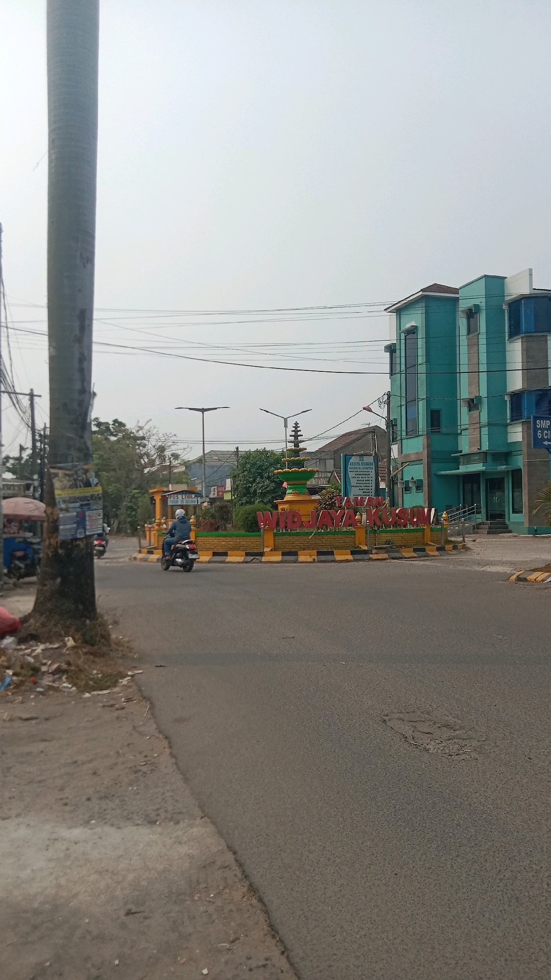 The image captures a view of a roundabout in Wanasari, Indonesia. A large, gray cylindrical pole stands prominently on the left side of the frame. The roundabout itself features a decorative monument with a tiered, golden structure topped by a finial, all set against a yellow and black striped border. Large red letters spelling "WIDJAYAKUSUMA" are visible in front of the monument. A blue and green building with multiple windows and vertical accents is situated to the right of the roundabout. A person is riding a motorcycle on the road within the roundabout. The road surface appears to be asphalt, with some patches of dirt and debris visible along the edge on the left. Power lines are strung across the sky. In the background, trees and smaller buildings can be seen. A sign for "SMP N 6 CI..." is partially visible on the right side of the blue and green building.