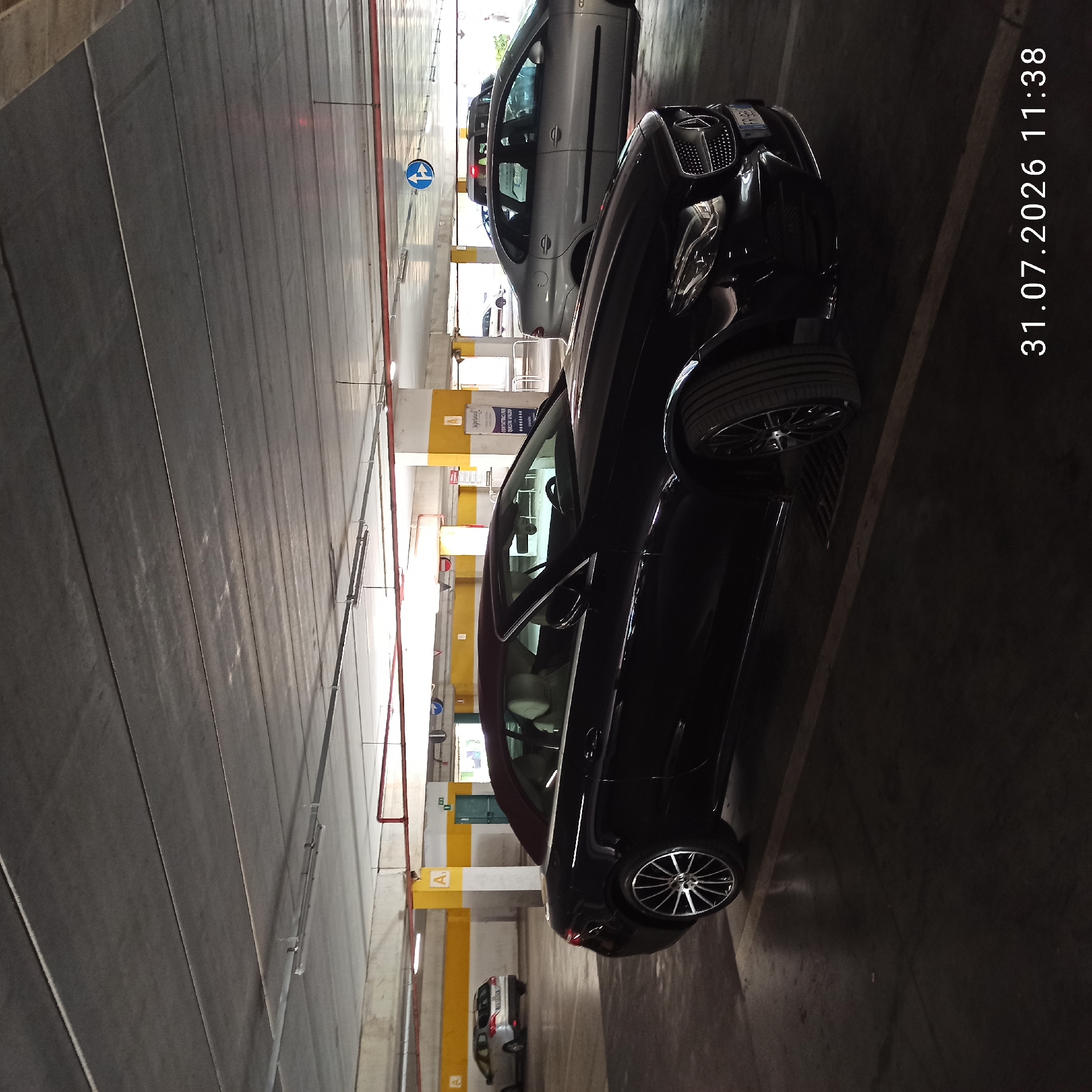 A black Mercedes convertible is parked in a multi-story parking garage. The garage has a concrete ceiling with exposed beams and lights, supported by concrete pillars painted yellow and white. The floor is concrete, with painted parking lines. In the background, a silver sedan is parked, and further back, another vehicle is visible. A sign with an arrow indicating left and straight turns is mounted on the ceiling. A timestamp in the bottom right corner reads "31.07.2026 11:38". The scene is consistent with a parking area, potentially at a shopping center or similar commercial establishment in Modugno, Italy.