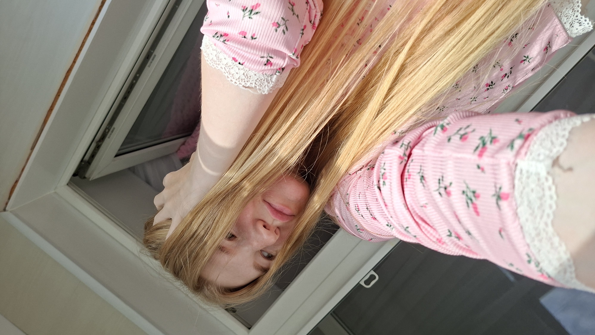An overhead, close-up, medium shot captures a young woman with long, blonde hair. She is indoors, by an open window, looking down and to the left at the camera. Her right hand is raised and resting on her head, with her fingers tucked into her hair. She is wearing a pink ribbed top with a small floral pattern of pink roses and green leaves. The top has white lace trim at the cuffs. The background consists of the white frame of the window and a sliver of the grey exterior, suggesting a balcony or outdoor space. The lighting is bright and natural, coming from above and to the right. The overall mood is casual and relaxed.