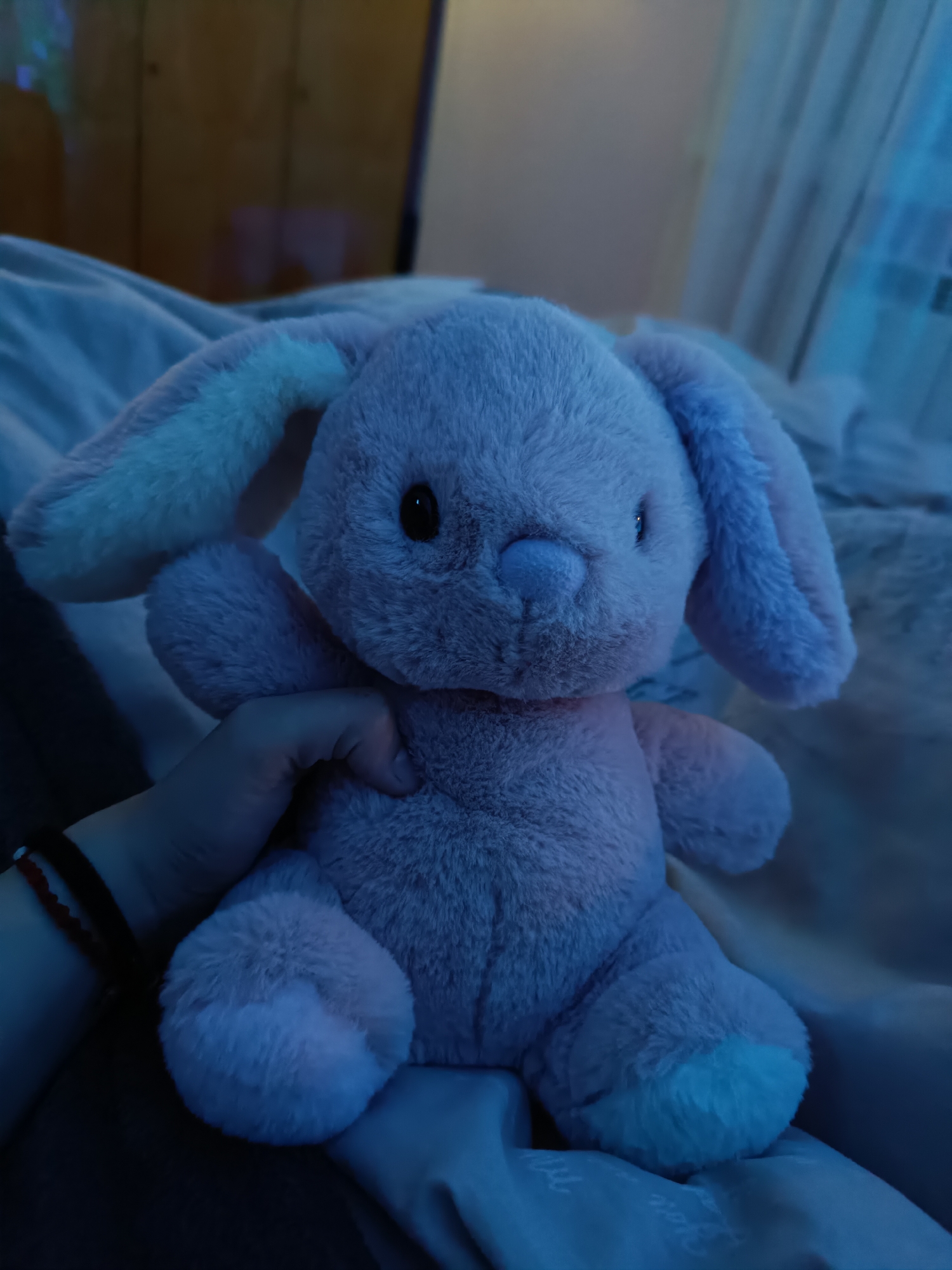 A pink stuffed bunny with black eyes is being held in a person's left hand. The bunny is positioned in the center of the frame. The background consists of bedding with blue light cast on it. In the far background, there is dark furniture. A bracelet is visible on the person's left wrist. The location is not identifiable as Cherkovitsa, Bulgaria based solely on the image.
