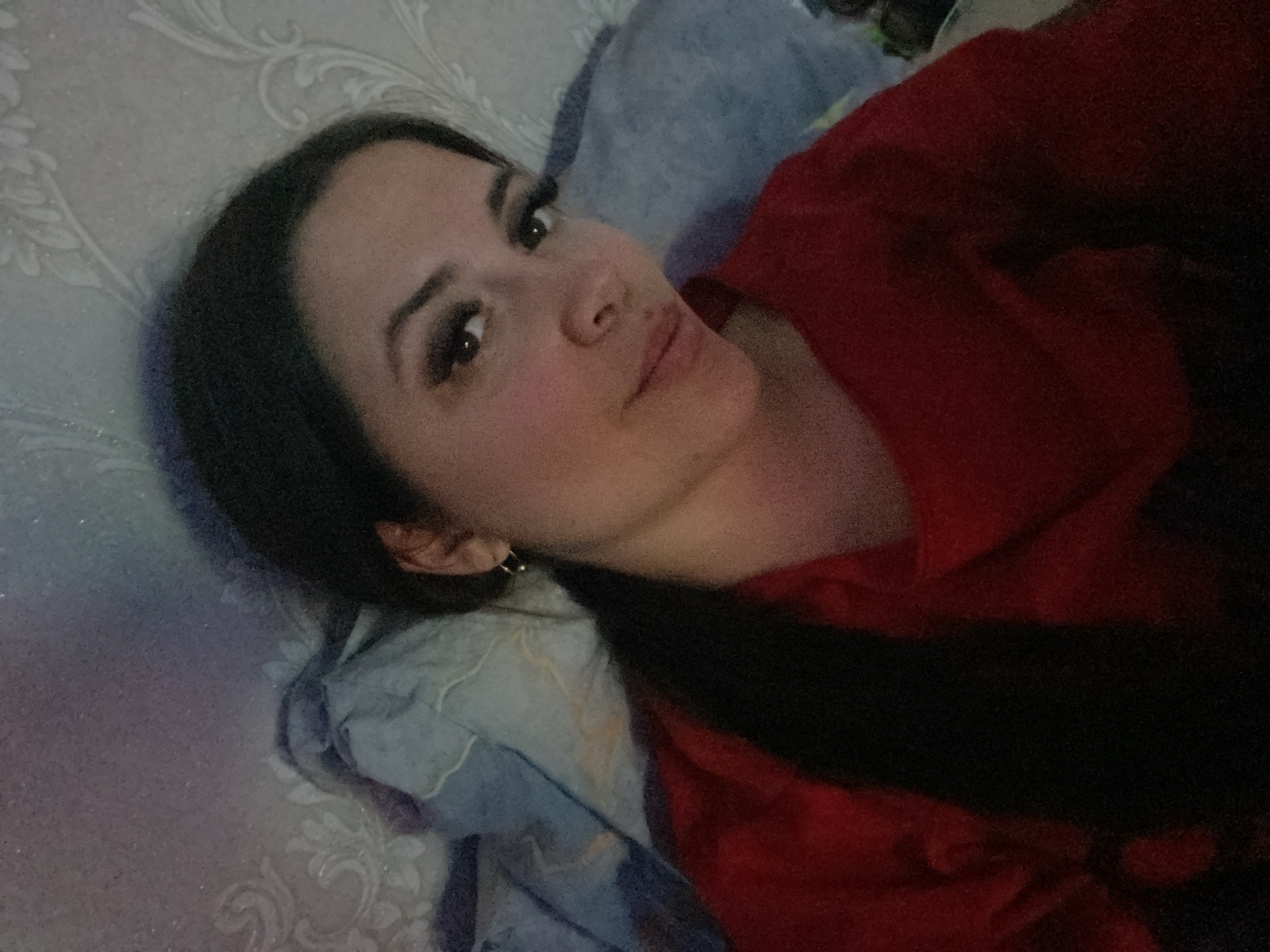 A young woman is lying down, her head resting on a blue pillow. She has long, dark hair, dark eyes, and wears an earring. She is wearing a red top. The background consists of a textured white surface, possibly wallpaper. The location is Chaikovsky, Russia.