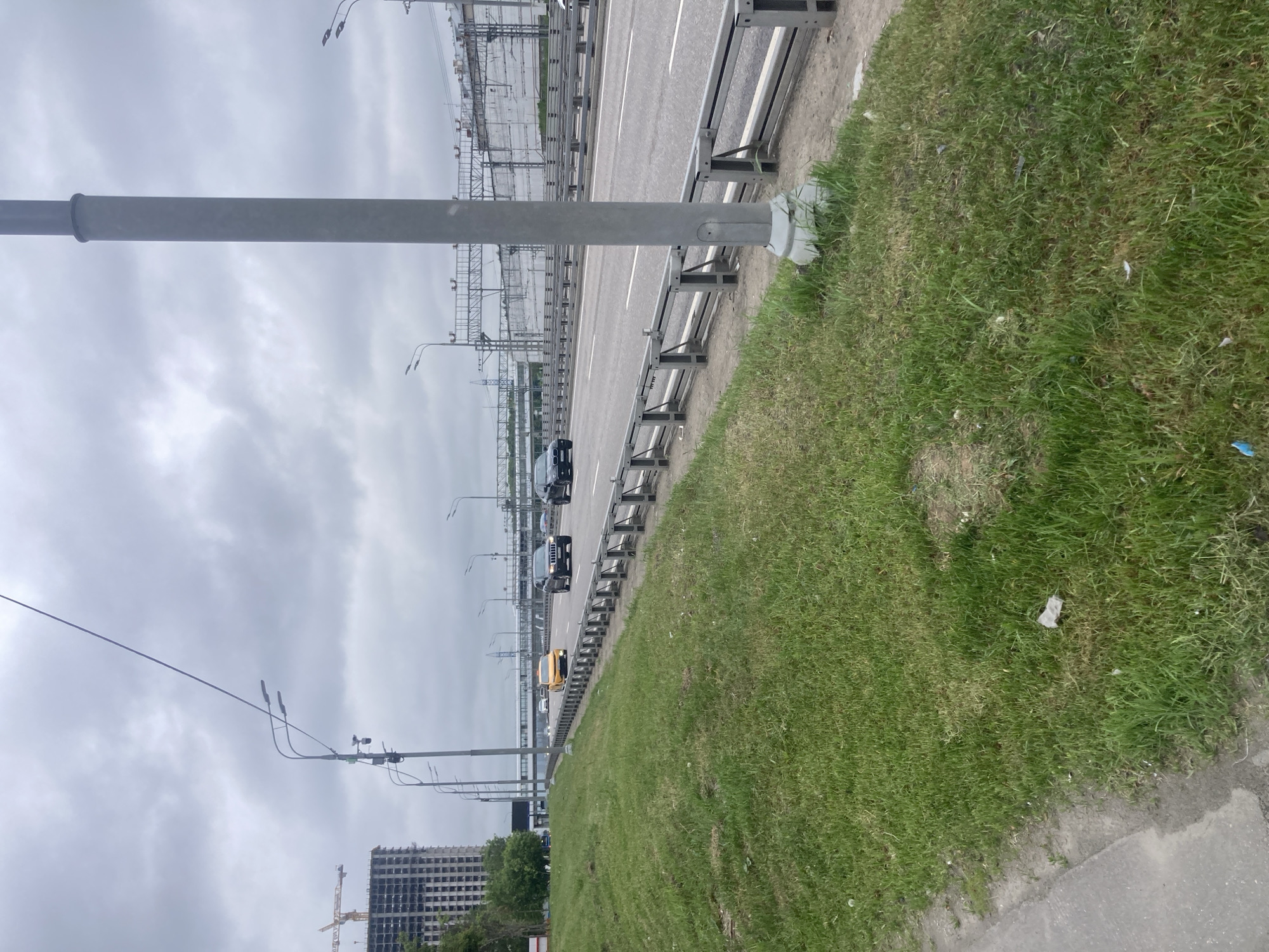 The image shows a section of a highway in Khimki, Russia, with a gray overcast sky above. The highway has multiple lanes and is lined with metal guardrails. Several vehicles are visible on the road, including cars and what appears to be a yellow taxi. A grassy embankment runs alongside the road, with a concrete sidewalk in the foreground. In the distance, there is a tall building under construction, with a yellow construction crane. Streetlights line the side of the road. A large gray pole is positioned in the foreground, appearing to be a support for overhead infrastructure. The overall scene is urban and depicts infrastructure typical of a highway environment.