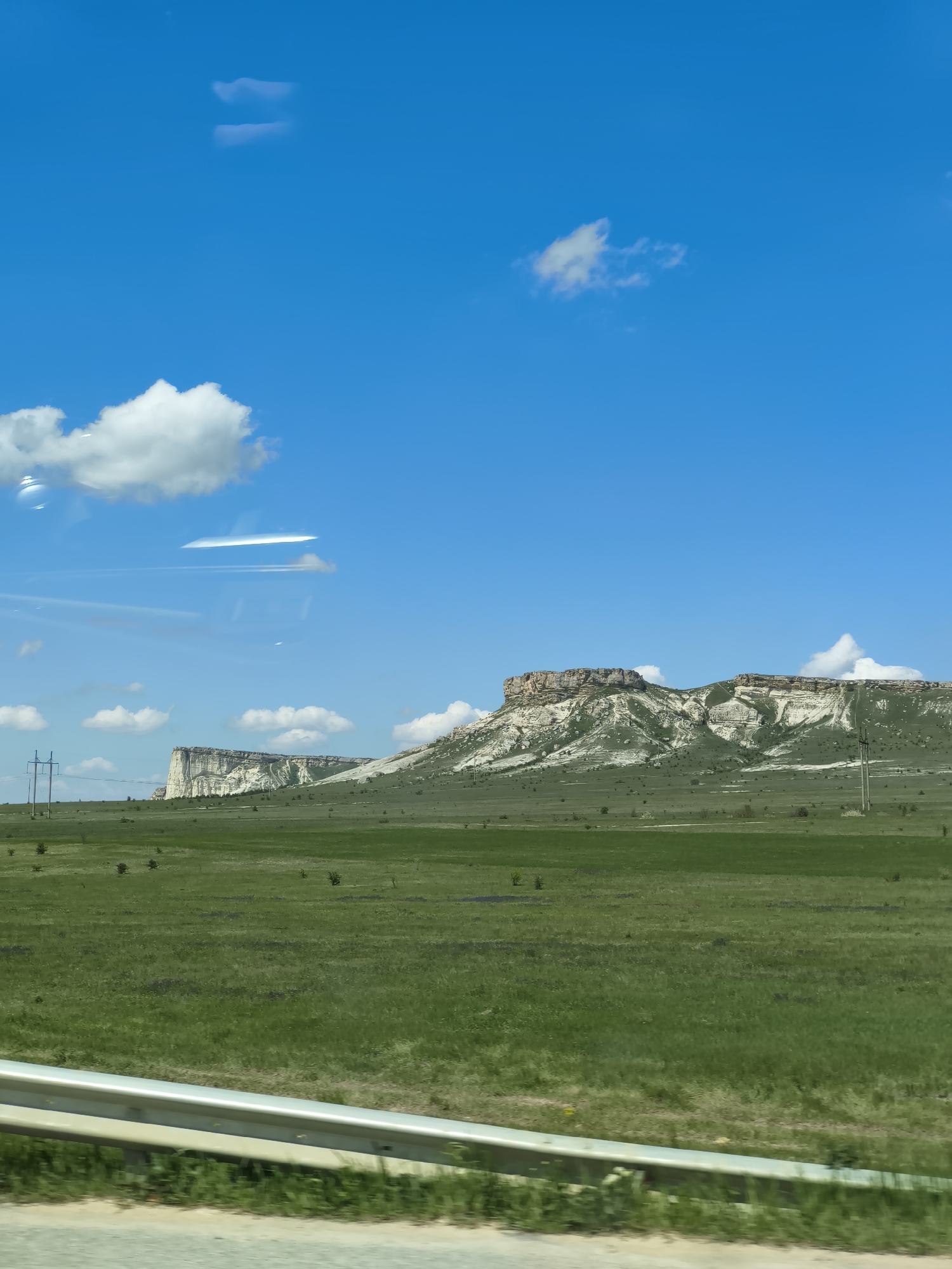 The image shows a landscape under a blue sky with scattered white clouds. In the distance, a plateau formation with white and brown layers is visible. The foreground is dominated by a green field, possibly grassland. A guardrail is visible at the bottom of the image. The location of the scene is Unknown, Unknown. There are no people in the image. The subjects are the landscape, the plateau formation, the field, and the sky. There are no actions or interactions visible.