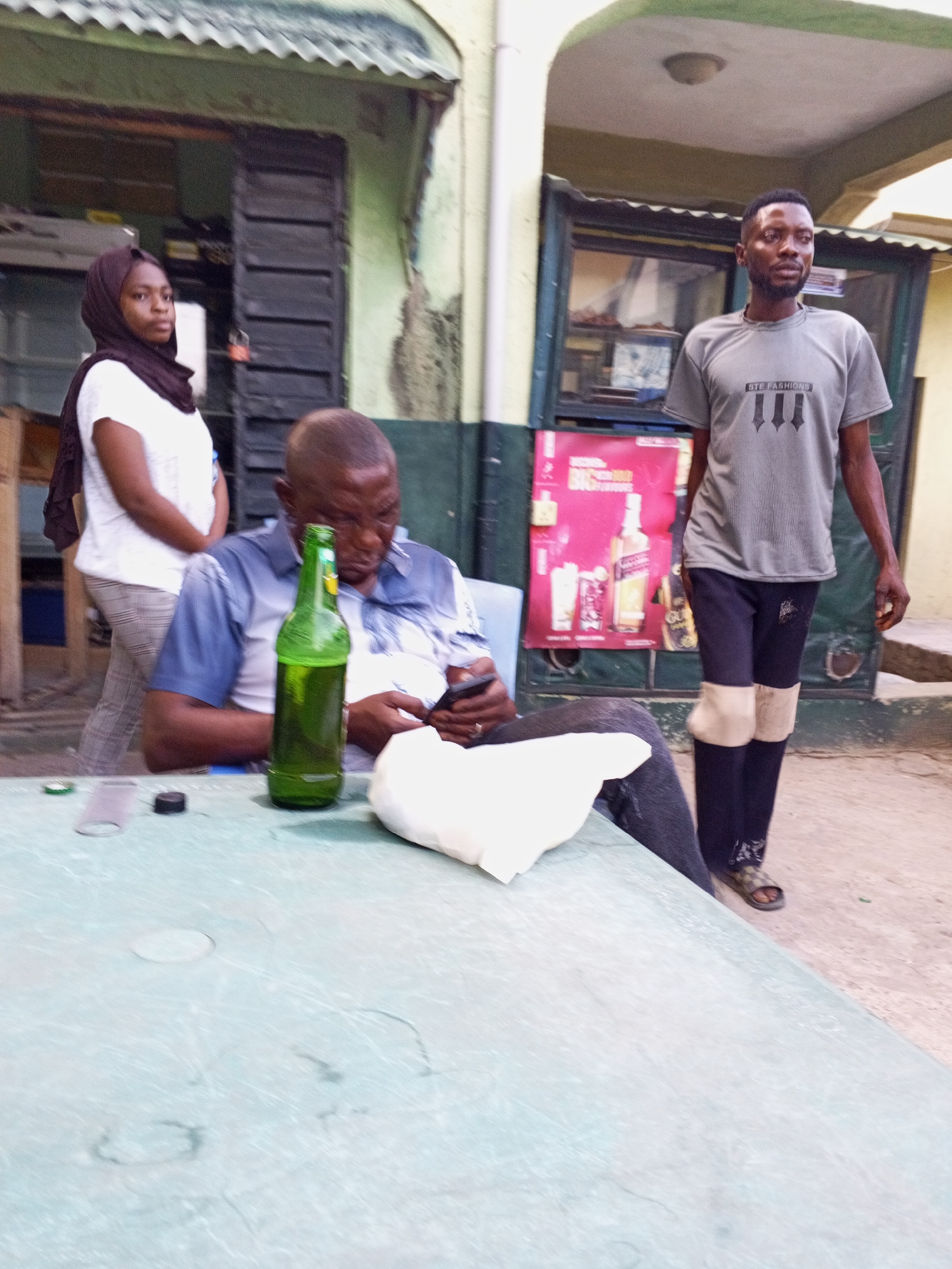 Zango-ndaji, Nigeria. A man wearing a blue, button-up shirt sits at a light-green table looking at his phone. A green bottle sits on the table next to him. A woman in a white shirt and headscarf stands behind him. A man wearing a grey shirt with the inscription “STE FASHIONS” stands on the right side of the image wearing black pants with beige patches. A poster is visible in the background behind the man.