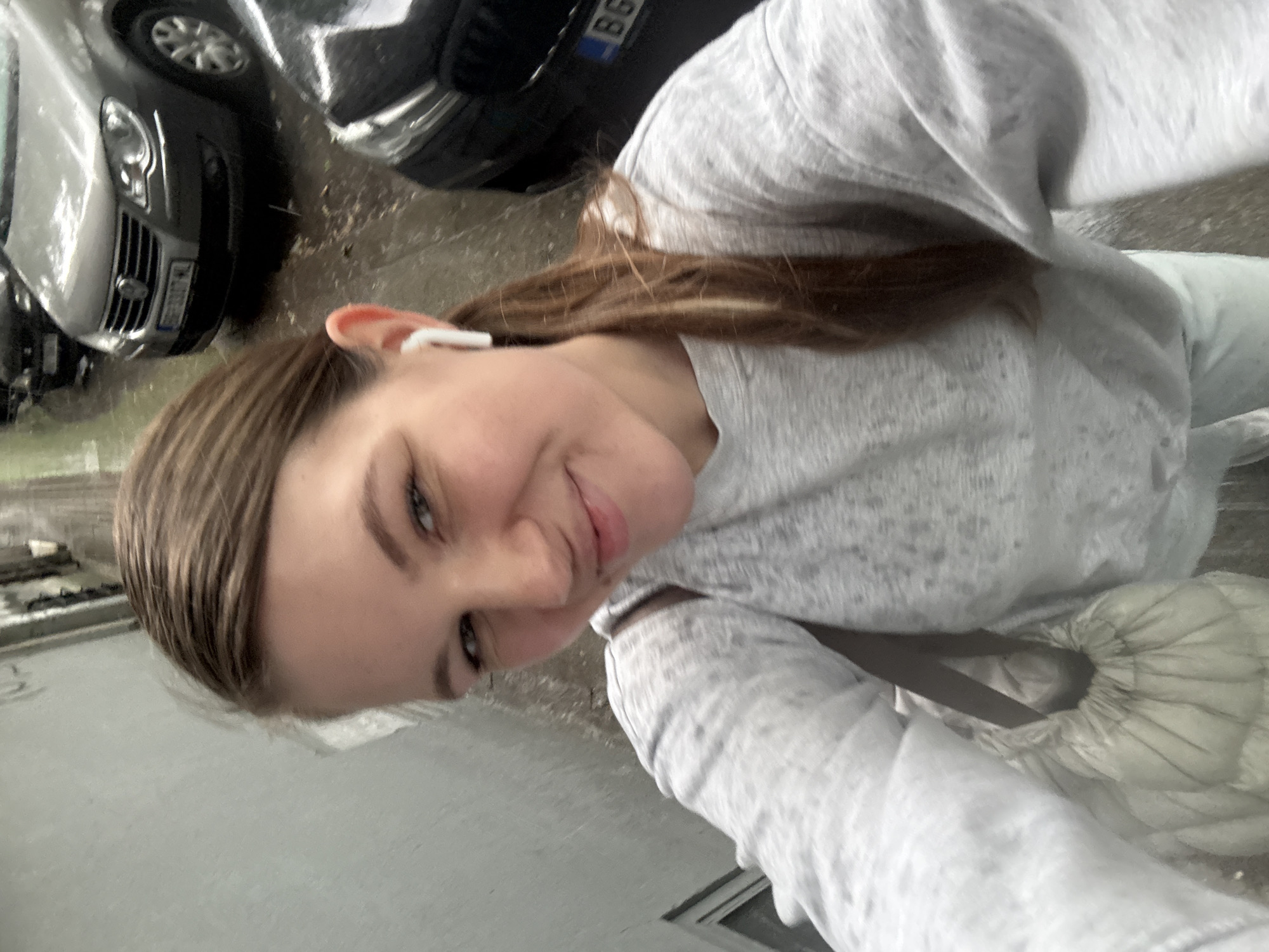 A young woman takes a selfie outdoors in Belgrade, Serbia. She wears a light-gray sweatshirt, and an earbud is visible in her left ear. Her hair is pulled back. In the blurred background, two cars are parked on the street. The license plate "BG" is visible on one. The ground is wet.