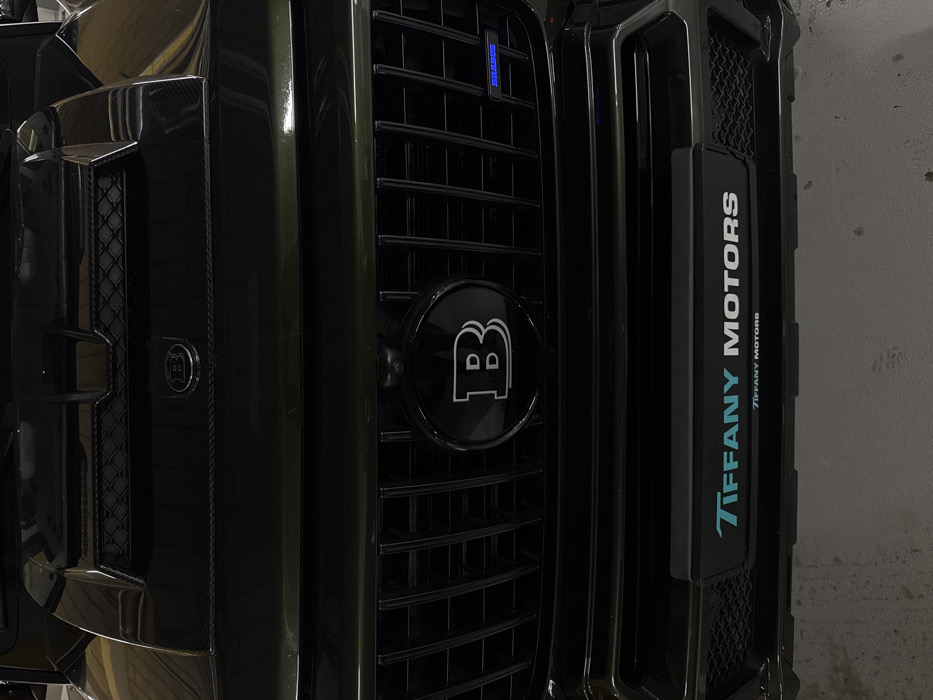 A close-up of a dark green Brabus SUV's front grille. The iconic "B" logo is prominently displayed on the center grille and smaller one on the left. A rectangular plate displaying "Tiffany Motors" in vertical text is visible on the right side. The grille consists of vertical bars with fine mesh inserts on either side. The overall impression is a luxurious and sporty vehicle.