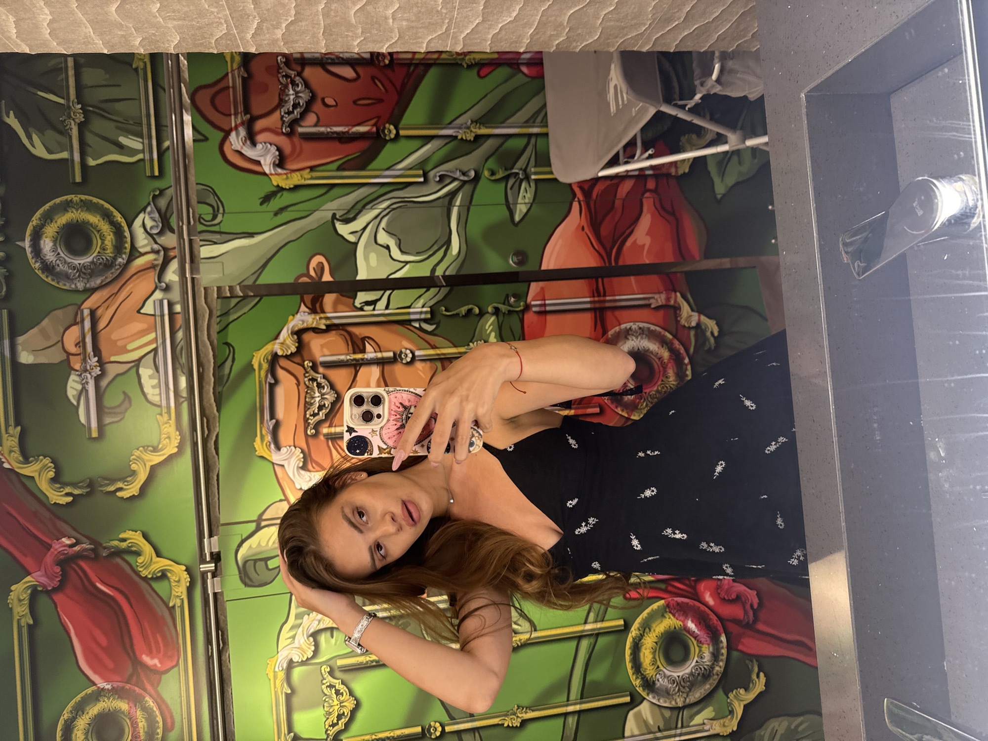 A woman with long brown hair takes a mirror selfie in a bathroom with a colorful mural. She is wearing a black dress with a floral pattern and jewelry on her left wrist. Her right hand is raised, holding a phone with a decorative case. The background features a vibrant mural with large red and orange floral motifs, green leaves, and metallic accents. A grey folding chair and a sink with a faucet are also visible. The location is specified as Plovdiv, Bulgaria, though no specific details of Plovdiv are evident in the image itself.