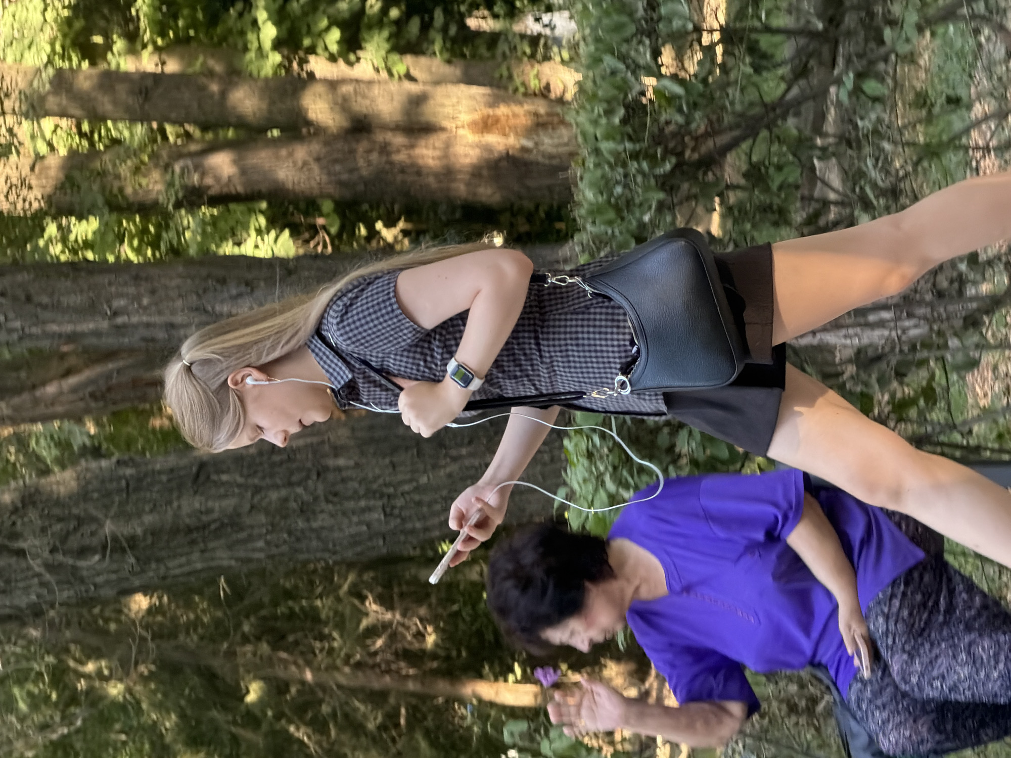 A young blonde woman with her hair in a ponytail walks in a park, holding a smartphone and wearing white earbuds. She is wearing a short-sleeved checkered shirt, a black skirt, and a black purse slung over her shoulder. A smartwatch is visible on her left wrist. To her left, an older woman with dark, curly hair is sitting on a bench, wearing a purple t-shirt and patterned pants. She is looking to her left and has her right hand raised. The background consists of trees and greenery, suggesting an outdoor, natural setting. The scene is captured in daylight.