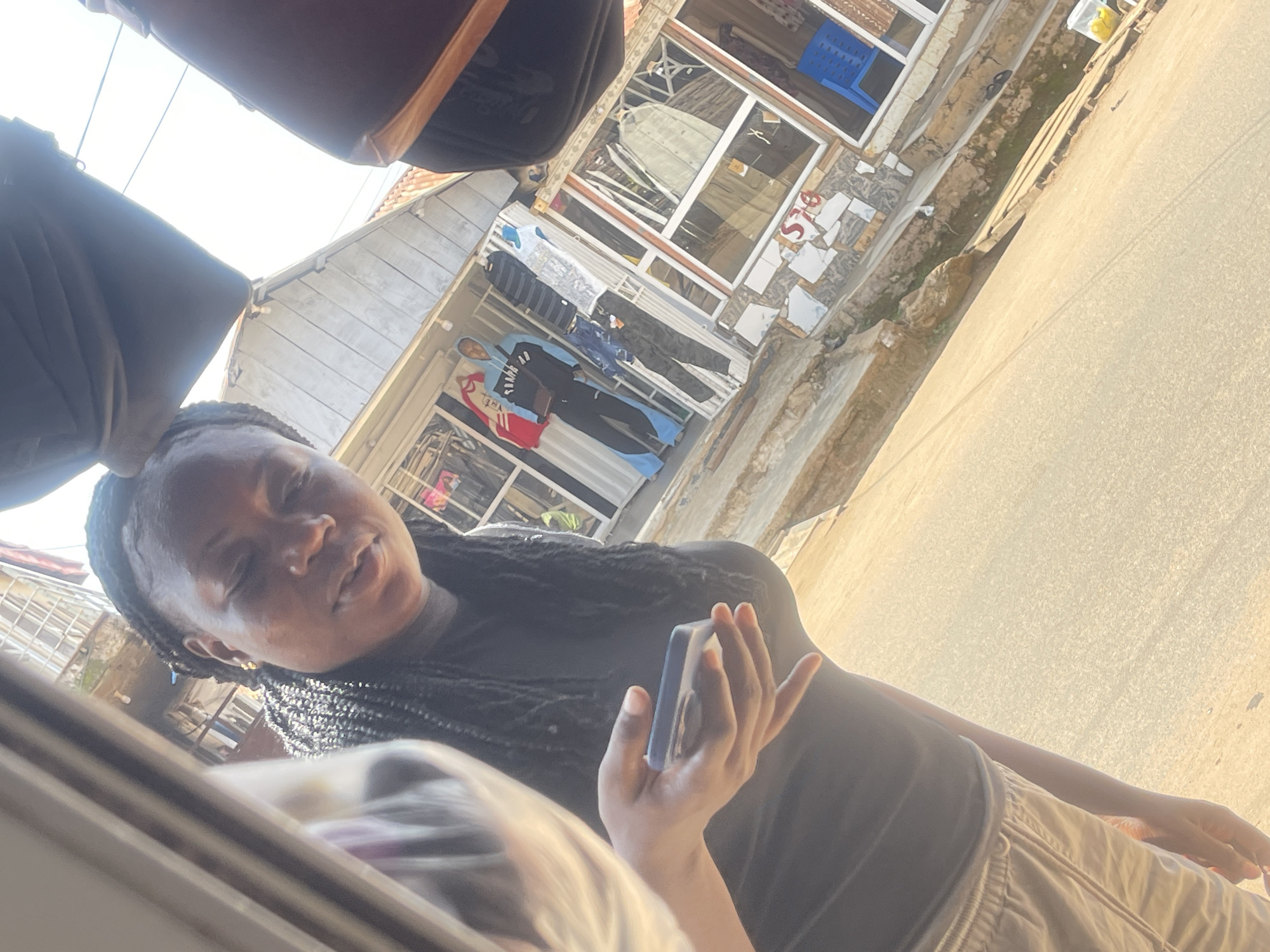 In this outdoor scene, a young Black woman with braided hair is holding a smartphone in her right hand and looking towards the camera. She is wearing a dark t-shirt and light-colored trousers. The background depicts a street in Abuja, Nigeria, with a shop displaying clothing on mannequins. The shop has a white facade with glass windows and various garments, including a blue and black tracksuit on one mannequin. To the right of the shop, a bright blue plastic chair is visible, and further to the right, a rack of clothing is seen. The ground is paved with concrete and asphalt, and there are some scattered rocks and debris. The overall lighting suggests it is daytime, with sunlight casting shadows.