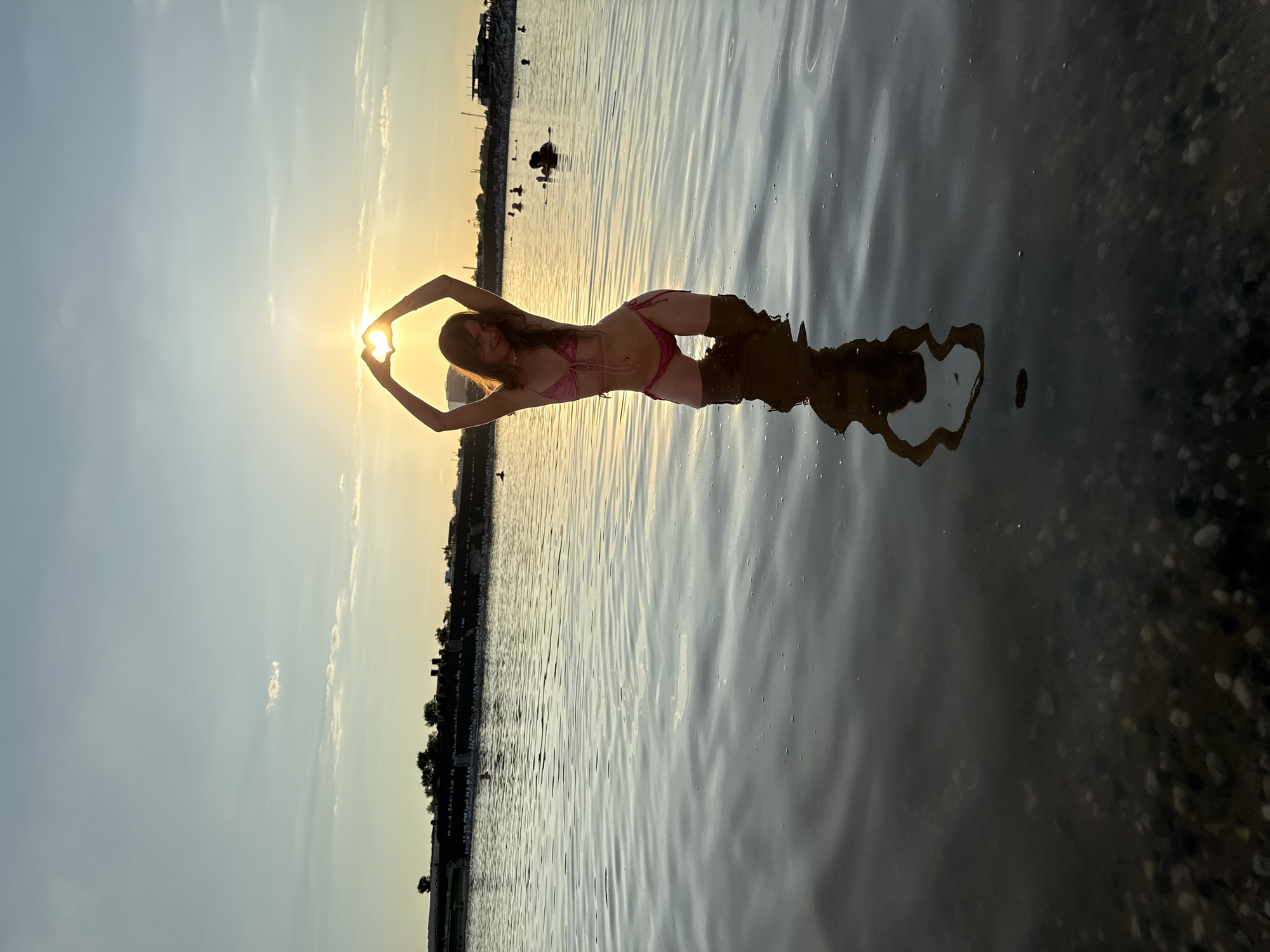 In the city of Sol-Iletsk, Russia, a young woman stands in the shallow water of a lake, forming a heart shape with her hands through the setting sun. She wears a pink bikini and her reflection is visible in the water. The sun is low in the sky, casting a warm glow and creating a lens flare through the heart shape. In the background, a distant shoreline is lined with buildings and trees, and other people can be seen in the water. The water's surface is rippled, and the lakebed is visible with pebbles in the foreground.