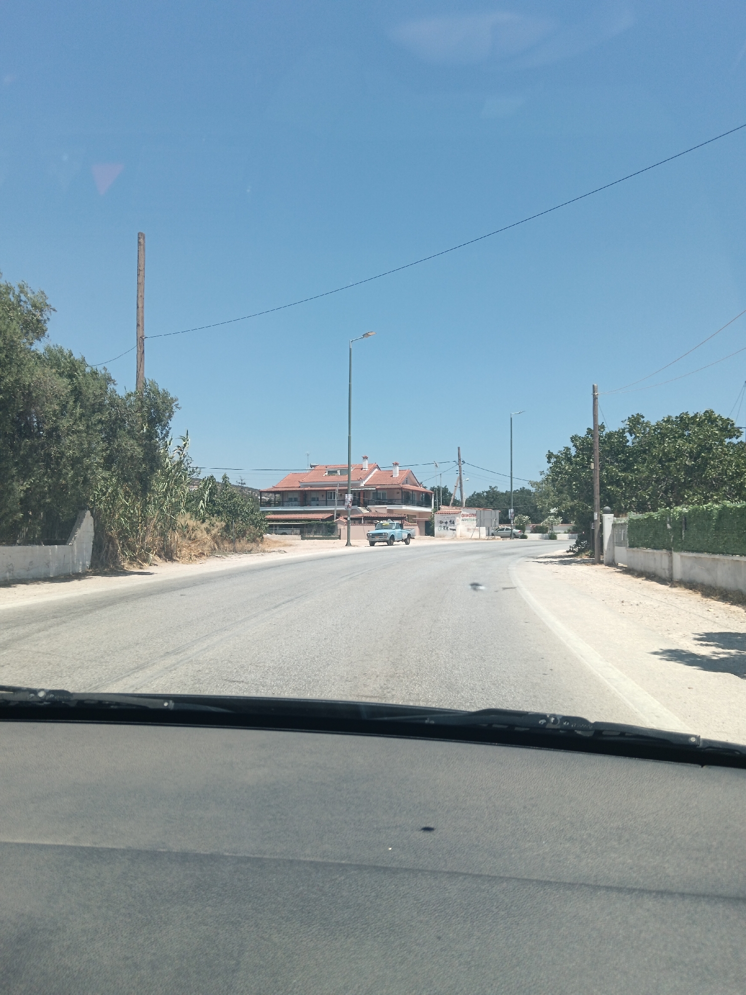 The image shows a view from inside a car driving on a road in Agia Marina, Greece. The road is paved and curves to the left. On the left side of the road, there are trees and a low white wall. On the right side of the road, there is a building with a red-tiled roof and a row of green bushes. There are also streetlights visible along the road. A light blue pickup truck is parked on the left side of the road in front of the building. The sky is clear and blue. The image appears to have been taken during the daytime.