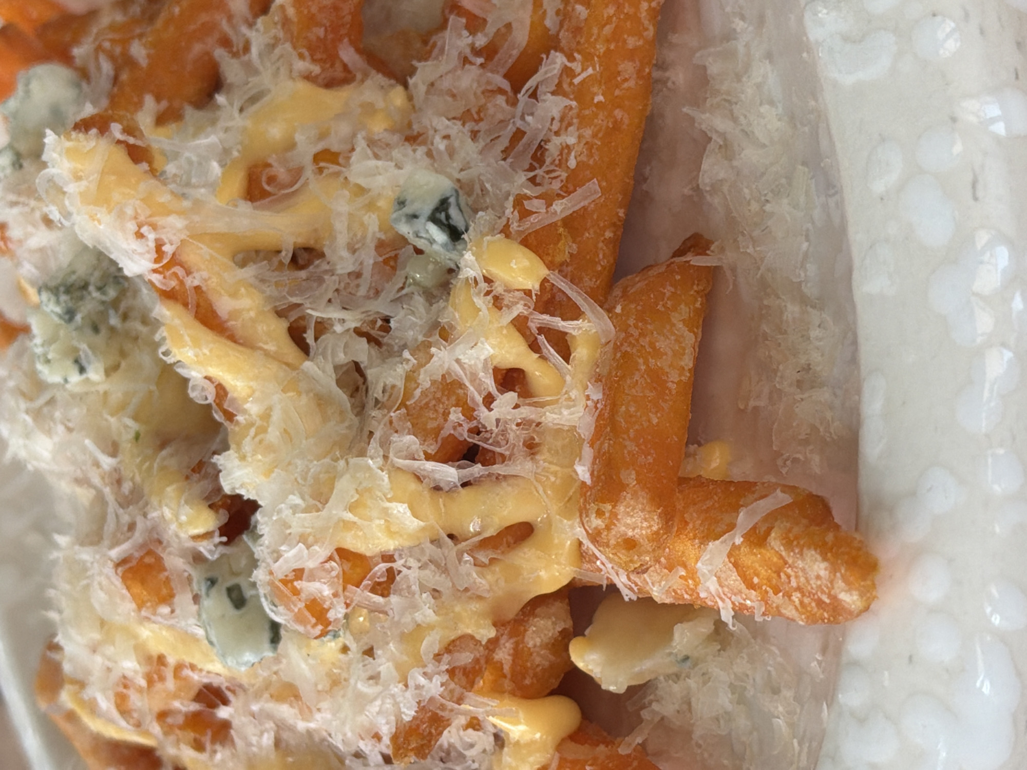 The image shows a close-up of sweet potato fries topped with melted cheese sauce, shredded cheese, and blue cheese crumbles. The fries are contained within a white, translucent container. The setting is not discernible, and there is no evidence to indicate a location in Moscow, Russia. No people are visible in the image.