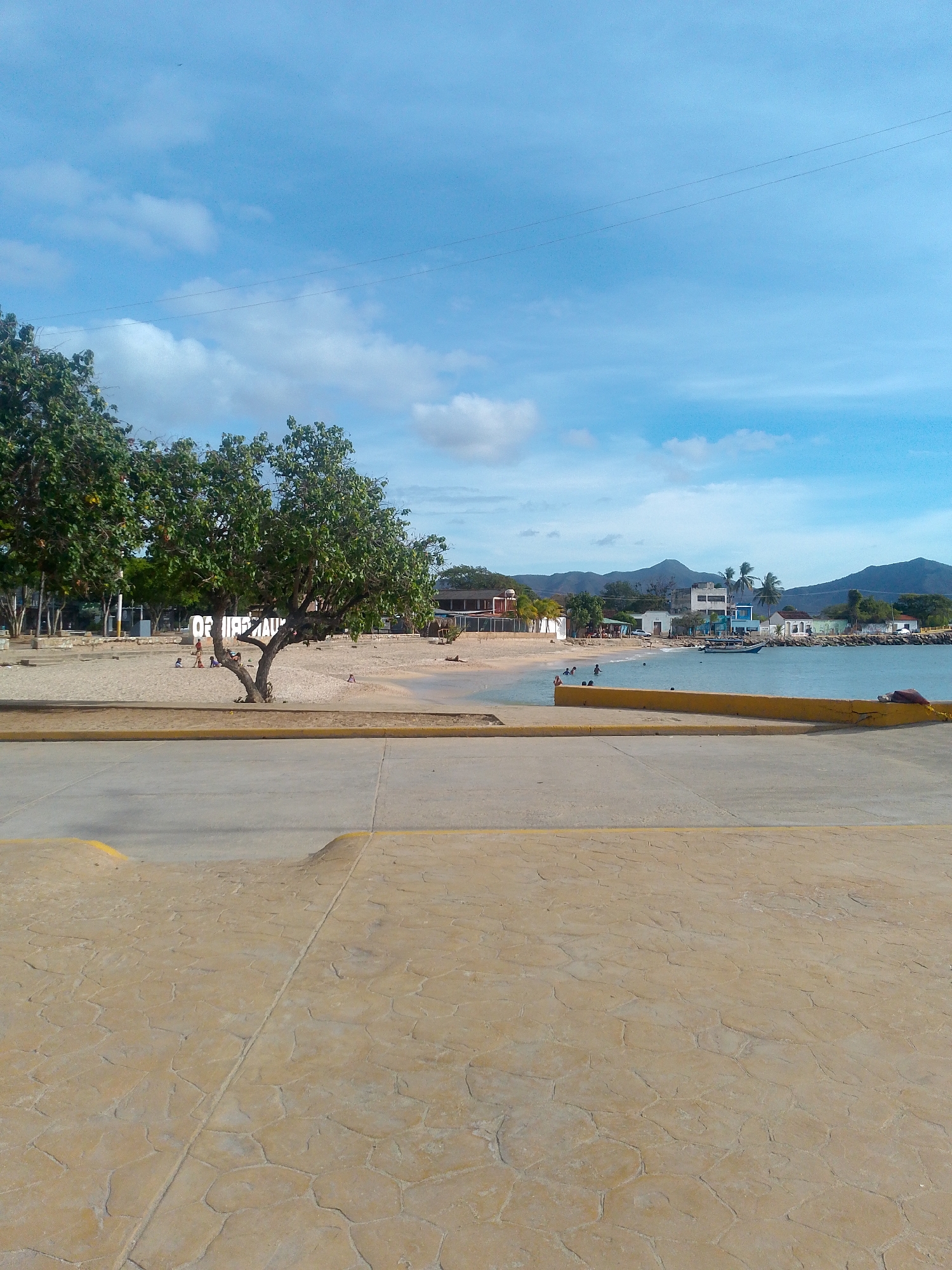 The image shows a coastal scene in Juan Griego, Venezuela. In the foreground is a paved area with a textured pattern, transitioning to a concrete road with a yellow painted edge. Beyond the road is a sandy beach area with sparse vegetation, including several trees. People can be seen on the beach, some near the trees and others closer to the water. In the background, a body of water with docked boats is visible. Buildings are situated along the waterfront, and hills rise in the far distance under a blue sky with scattered clouds. An overhead wire stretches across the sky. A large white sign sits on the beach among the trees.