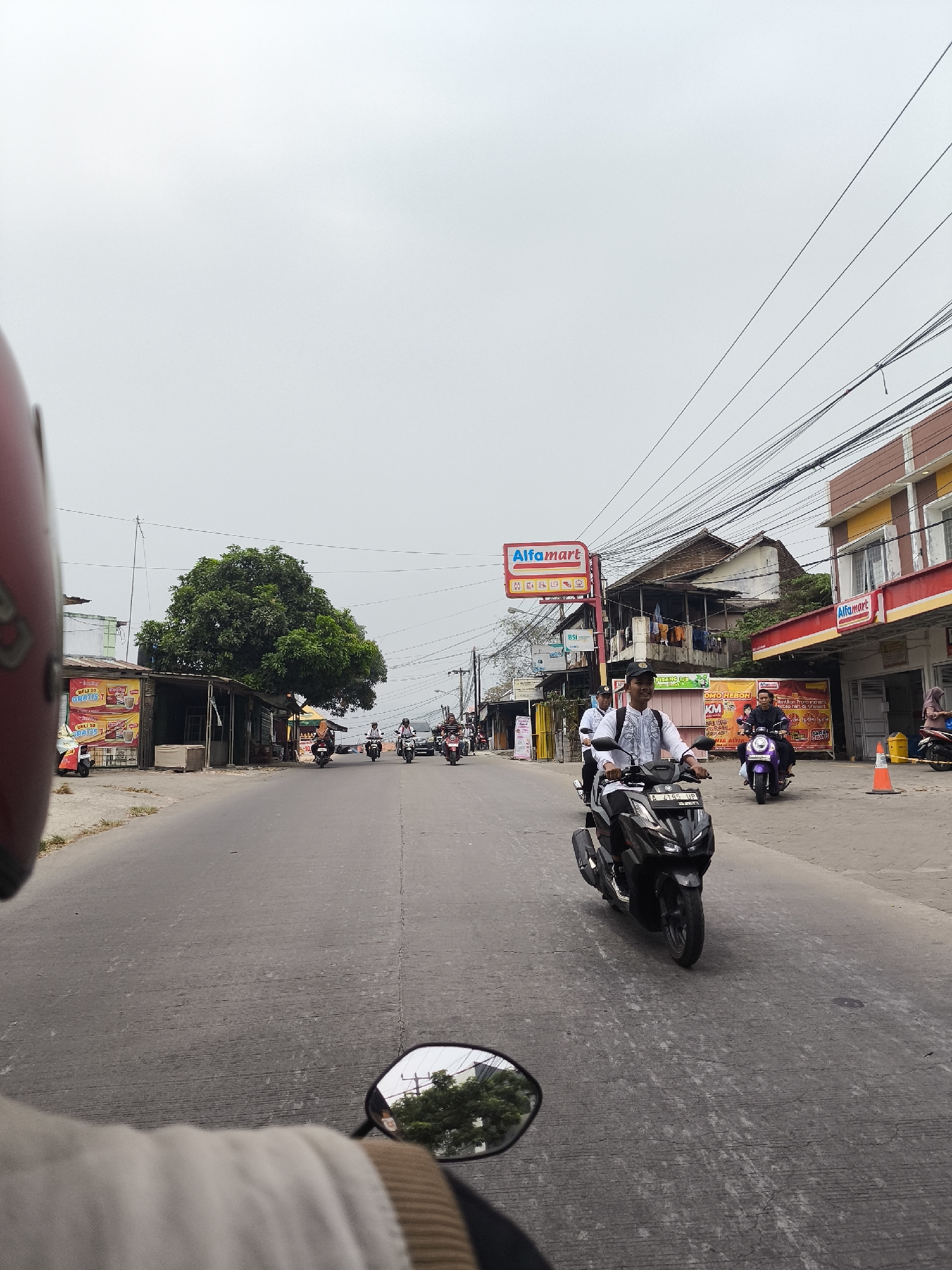 The image depicts a street scene in Cilegon, Indonesia, on an overcast day. The perspective is from the viewpoint of someone riding a motorcycle, as evidenced by the reflection of a rearview mirror showing trees and a building, and a portion of the rider's shoulder and the back of a red helmet in the foreground. The road is paved and appears to be a two-lane street with moderate traffic.
Several people on motorcycles are visible. In the foreground, a person wearing a white shirt and a dark jacket is riding a black motorcycle. Further down the road, another individual is on a purple scooter. Other motorcycles and riders are scattered throughout the scene.
Along the right side of the road are commercial buildings. An Alfamart store with its distinctive red and white signage is prominent. Adjacent to it is another building, possibly a shop or residential unit, with a brown and yellow facade. Wires are strung overhead, typical of urban infrastructure.
On the left side of the road, there are smaller, older-looking structures with tin roofs and open fronts, suggesting local small businesses or stalls. A large, leafy green tree stands between these structures and the road. Advertising banners are visible on some of the buildings. An orange traffic cone is placed on the sidewalk near the Alfamart. The overall impression is a bustling local street with everyday commerce and transportation.