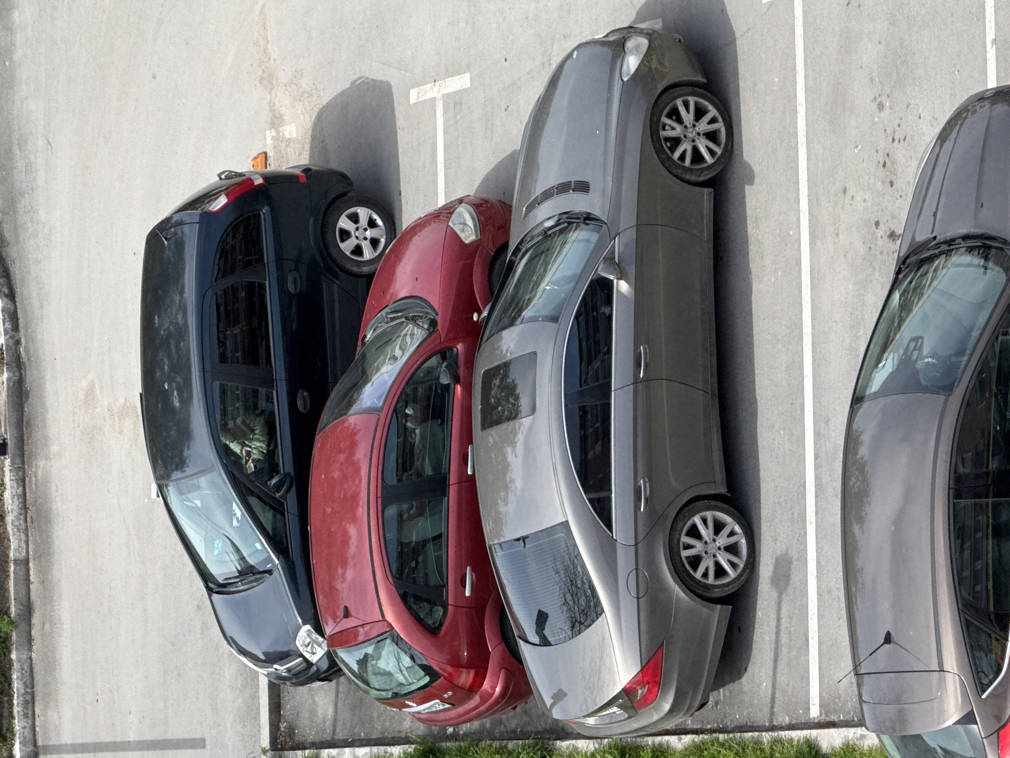 The image presents an aerial view of a parking lot in Varna, Bulgaria. Four cars are parked in a row. From left to right, the cars appear to be dark blue, red, gray, and dark gray. The parking spaces are marked with white lines. The surface of the parking lot is gray concrete or asphalt. Vegetation is seen in the lower left and right corners of the image. The parking lot is near a street or sidewalk.