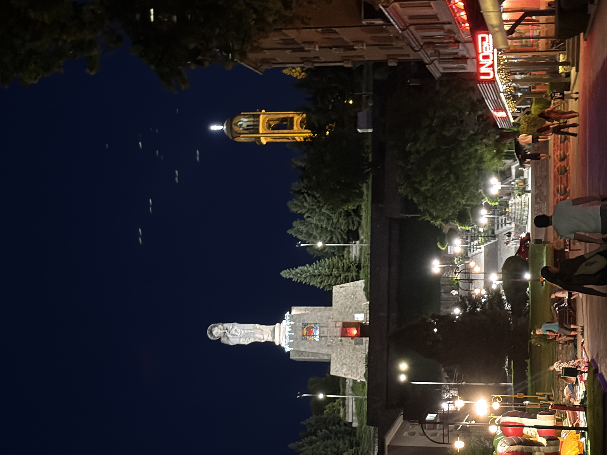 At night in Haskovo, Bulgaria, the "Eternal Fire" monument and the city's bell tower are illuminated. The monument, a statue of Mary with child, stands tall on a stone pedestal, adorned with festive lights and a stained-glass window. A red light emanates from an opening at the base of the monument. The bell tower, a striking yellow structure, is lit from within and topped with a cross. In the foreground, a vibrant street scene unfolds. Families stroll along a path lined with illuminated street lamps and trees. An inflatable playground with bright colors is visible on the left, while on the right, the "UNO" restaurant glows with neon lights. The sky above is a deep, dark blue, with faint streaks of light, possibly from passing aircraft. The overall atmosphere is one of a lively evening in a Bulgarian city.
