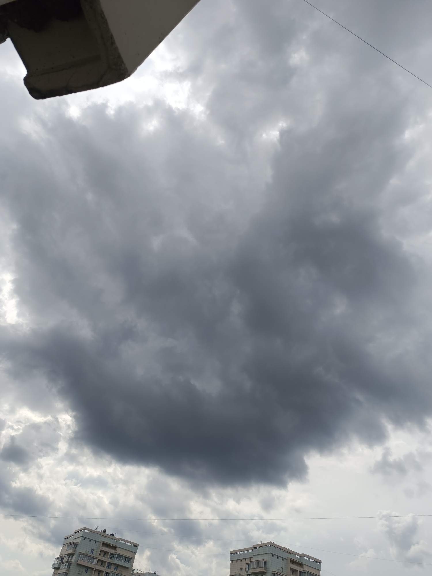 The image shows a cloudy sky above two apartment buildings. The clouds are dark and heavy, suggesting an approaching storm. The buildings are modern and appear to be in good condition. There are no people visible in the image. The scene is set in a city, but the specific location is unknown.
