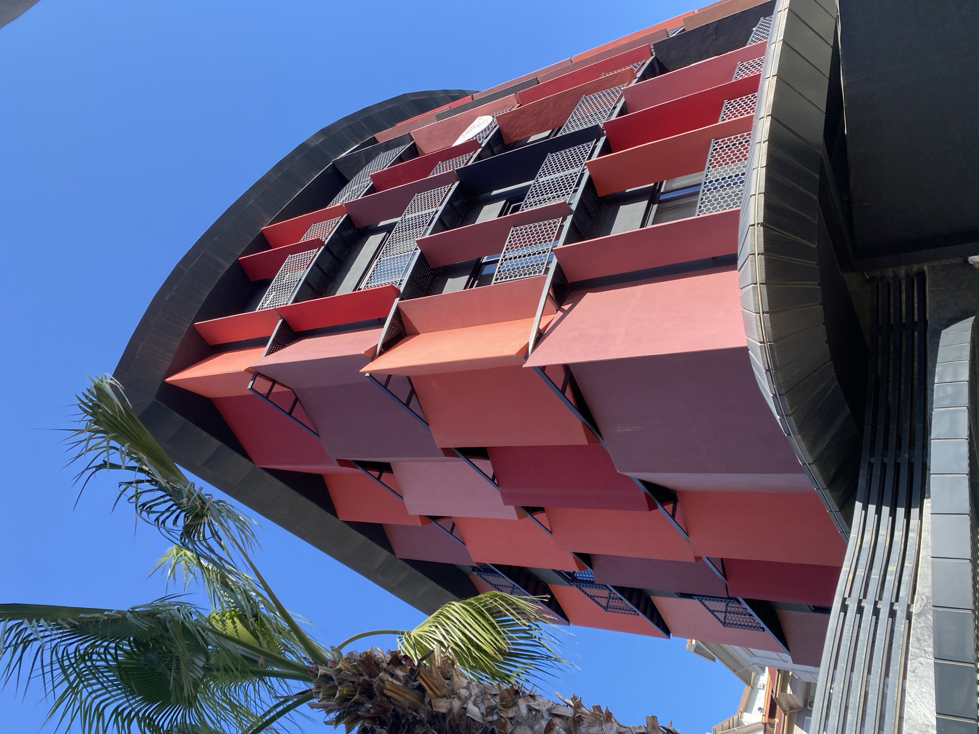 This image presents a low-angle view of a modern building against a clear, vibrant blue sky. The main subject is the building's distinctive facade, which is composed of numerous rectangular panels arranged in a tessellated pattern. These panels display a gradient of colors, ranging from various shades of red and maroon to peach and salmon, creating a dynamic visual effect. The panels appear to be angled outwards, possibly functioning as sun shades or balcony fronts, with metallic, perforated screens featuring a honeycomb pattern visible behind them and in windows.
The upper portion of the building and its curved roofline are dark grey or black, providing a strong contrast to the colorful facade. On the right side, an adjacent part of the structure or another building is also depicted in dark tones with vertical design elements. In the lower-left foreground, a green palm tree with a textured brown trunk extends into the frame, its fronds adding a natural touch to the architectural scene.
The bright sunlight suggests it is daytime, likely midday or early afternoon, casting sharp shadows that enhance the building's three-dimensional qualities. The weather is clear and sunny, indicated by the unblemished blue sky. There is no discernible activity taking place, and no visible text can be found in the image. The scene conveys a sense of contemporary architecture in a bright, Mediterranean-like setting, consistent with Alanya, Turkey.