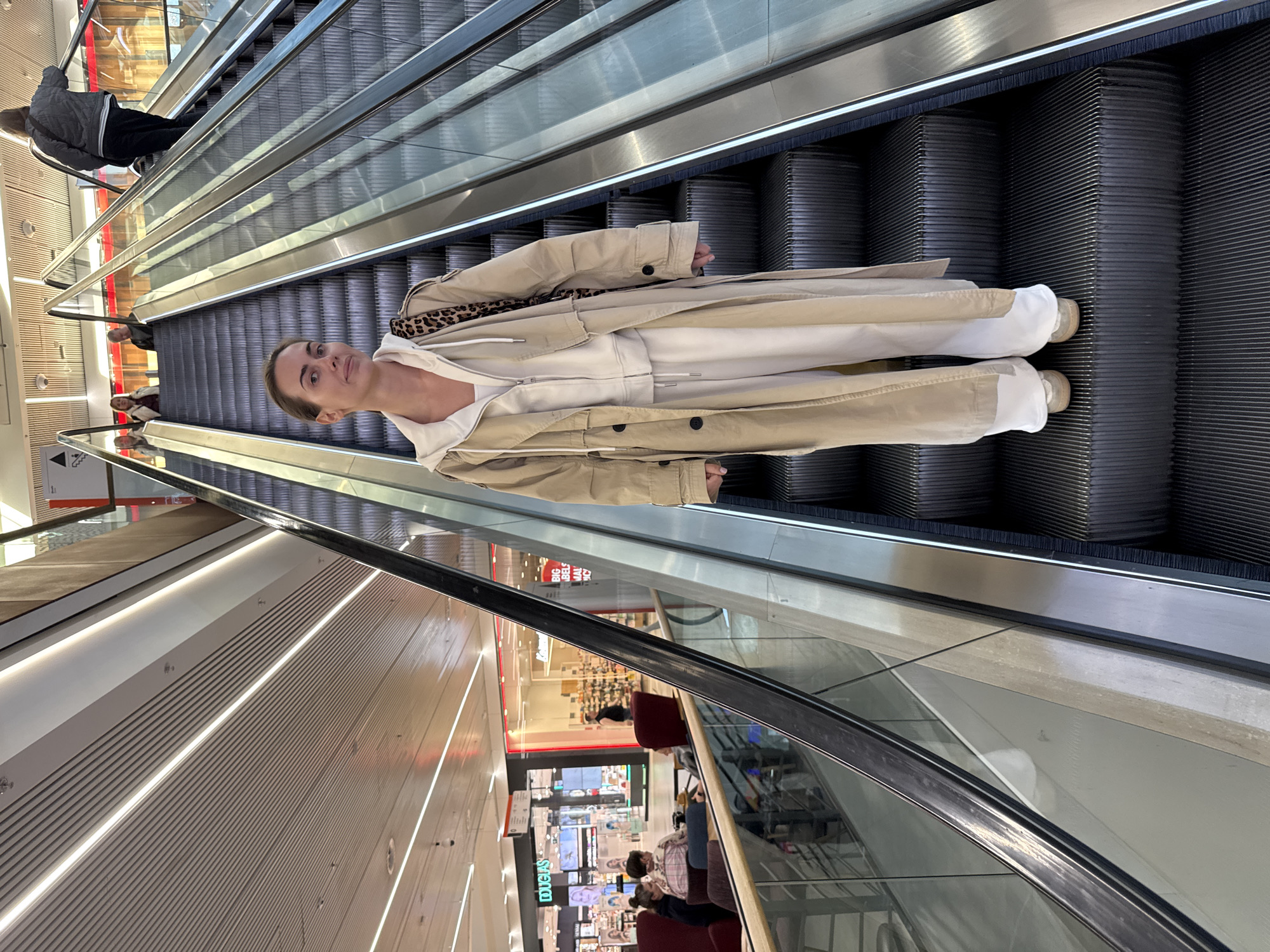 A woman is depicted standing on an escalator inside a modern shopping mall. She occupies the central-right portion of the frame, posing for the camera with a neutral expression. She has light brown hair pulled back and is dressed in a light-colored hooded top, a tan trench coat, and light-colored trousers, with matching light-colored shoes. A leopard print pattern, possibly a scarf or the coat's lining, is visible around her neck.
The escalator features black, ribbed steps and clear glass balustrades with metallic handrails. It appears to be ascending. Other individuals are present on the escalator; one person in a dark jacket and pants is visible further up, and more people are seen in the reflections of the mall's interior. The setting is brightly illuminated by artificial indoor lighting, creating a clean and contemporary ambiance.
Reflections on the glass surfaces and polished floors reveal various elements of the shopping center, including storefronts, display cases, and other patrons. Visible text includes "BIG LABELS" on a red sign and "DOUGLAS" on a store sign, both appearing in background reflections. A small directional sign with an arrow and a person icon is also faintly visible at the bottom. The image is presented with a vertical orientation, making the escalator appear to run diagonally upwards. The location context is Warsaw, Poland.