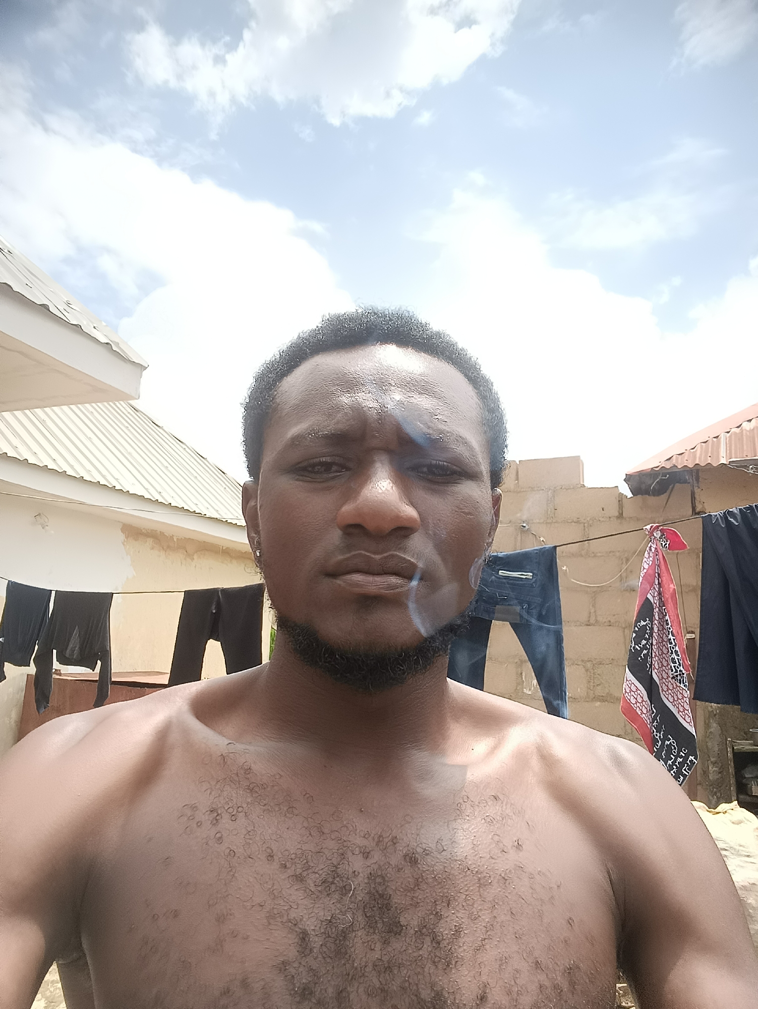 A close-up, waist-up, outdoor selfie shows a shirtless Black man exhaling smoke from his mouth. He has short, dark, curly hair, a full beard, and a small earring in his left ear. His shoulders and chest are visible, with a light covering of dark hair on his chest. The background features a brick wall, laundry hanging on a clothesline, and a portion of a corrugated metal roof. The sky above is blue with white clouds. The scene is consistent with a residential outdoor setting in Jos, Nigeria, possibly a backyard or an open space where laundry is hung to dry.
