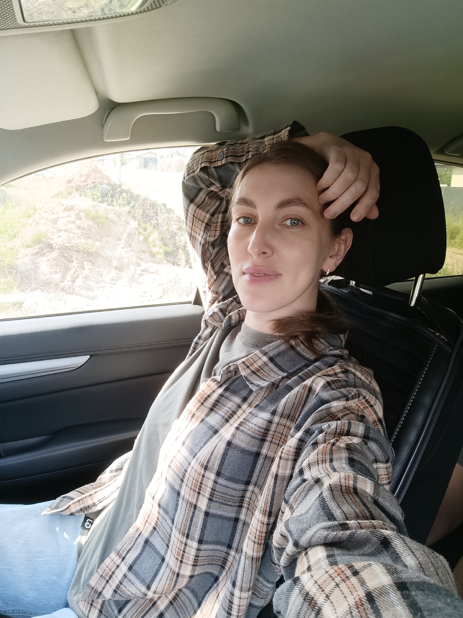A young woman with light brown hair and blue eyes is seated in the passenger seat of a car. She is wearing a grey, tan, and white plaid shirt over a green t-shirt, and light blue jeans. Her right arm is resting on her head, and her hand is touching her forehead. She is looking towards the camera with a slight smile. The interior of the car, including the headrest, door panel, and overhead handle, is visible. Through the car window, a sunlit, rural landscape with trees and vegetation can be seen. The time of day appears to be daytime, with bright sunlight casting shadows. The woman's expression suggests a relaxed and content mood. The scene is likely set in Chaikovsky, Russia, given the provided location context.