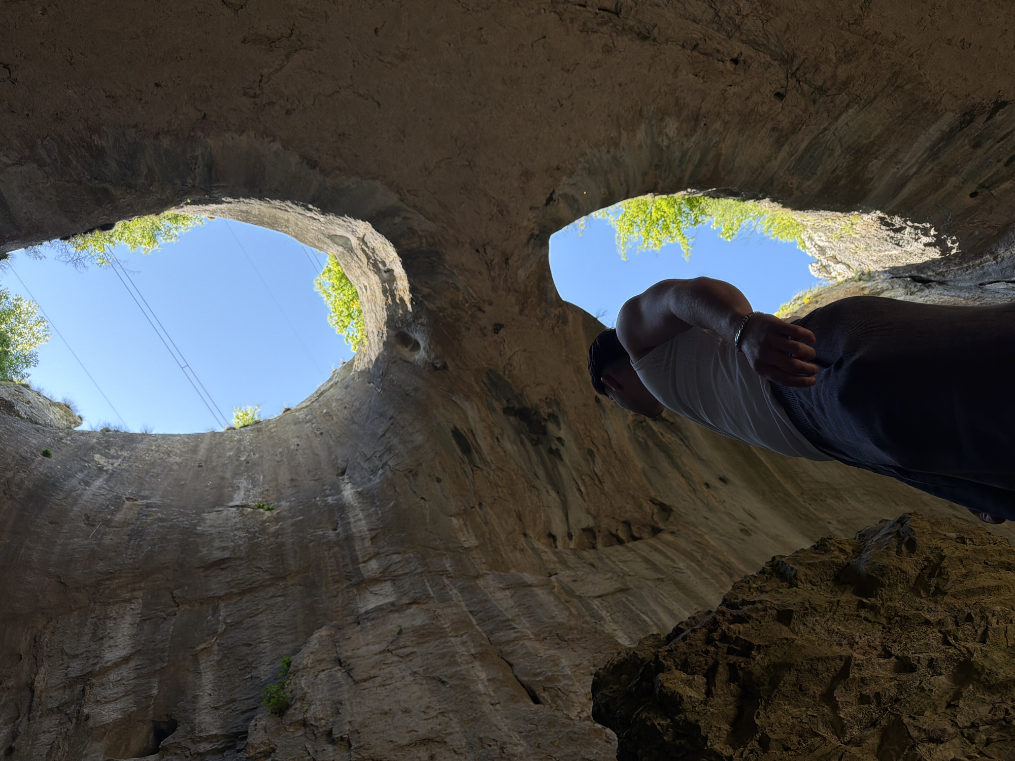 The image captures a low-angle view from within a cave system, identified as Prohodna Cave near Karlukovo, Bulgaria. The cavern features two large, symmetrical openings in the ceiling, revealing a bright blue sky and sparse greenery. A person is positioned along the right edge of the frame, leaning into the shot, wearing a white top and dark pants. The cavern walls are textured with visible rock layers and variations in color, ranging from light to dark gray. The foreground includes rocky terrain, indicating the cave's natural environment. Electrical cables are visible across the holes to the sky.