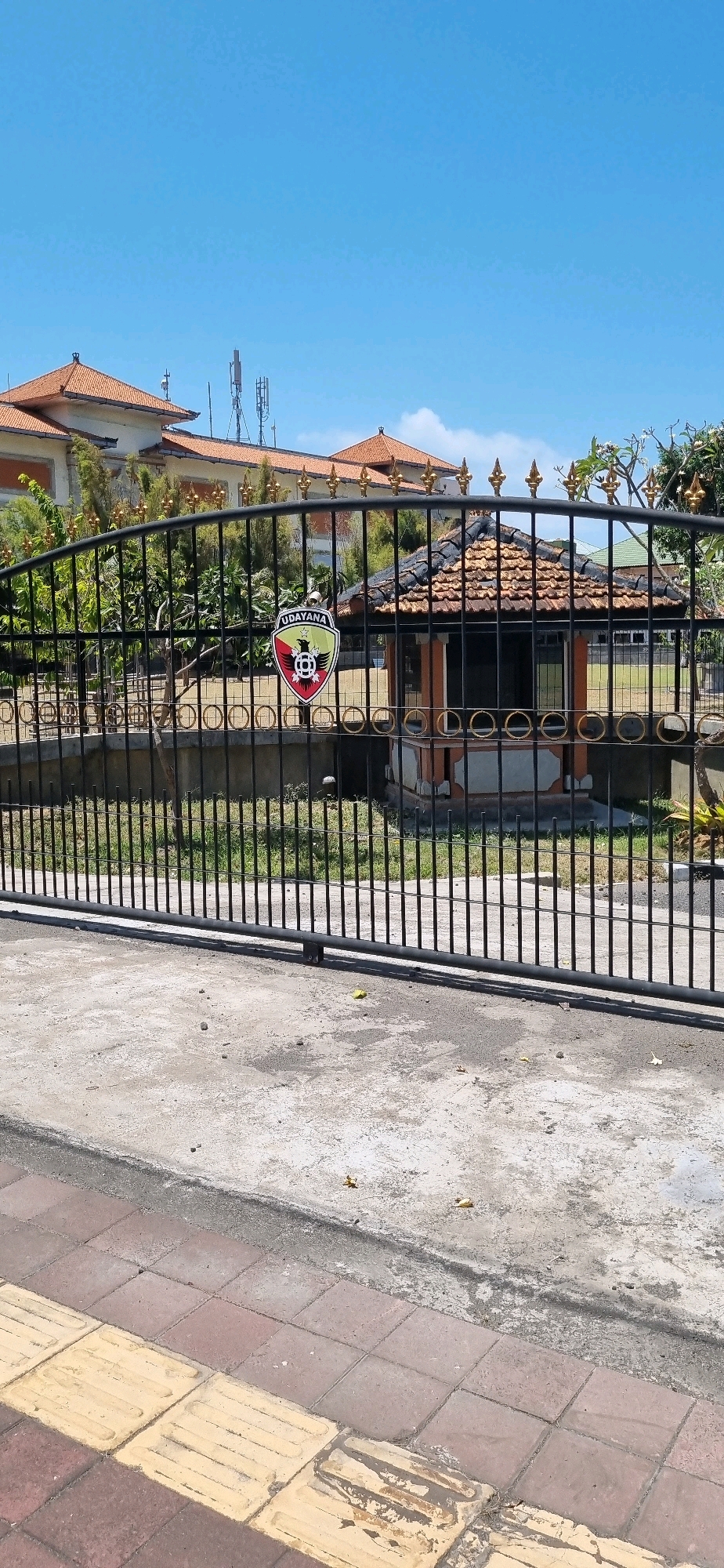 This is a ground-level, eye-level shot of a black metal gate with gold decorative accents. The gate is in front of a building with a red tile roof and a logo on it that says "UDAYANA". There is a small, octagonal structure with a red tile roof behind the gate. The ground in front of the gate is made of concrete, and there is a sidewalk with yellow tactile paving stones. The sky is blue and clear with some clouds. The image appears to have been taken during the day, likely in the morning or afternoon due to the bright sunlight and clear sky. This location is likely a government building or educational institution in Denpasar, Indonesia, as "Udayana" is the name of a prominent university in Denpasar.