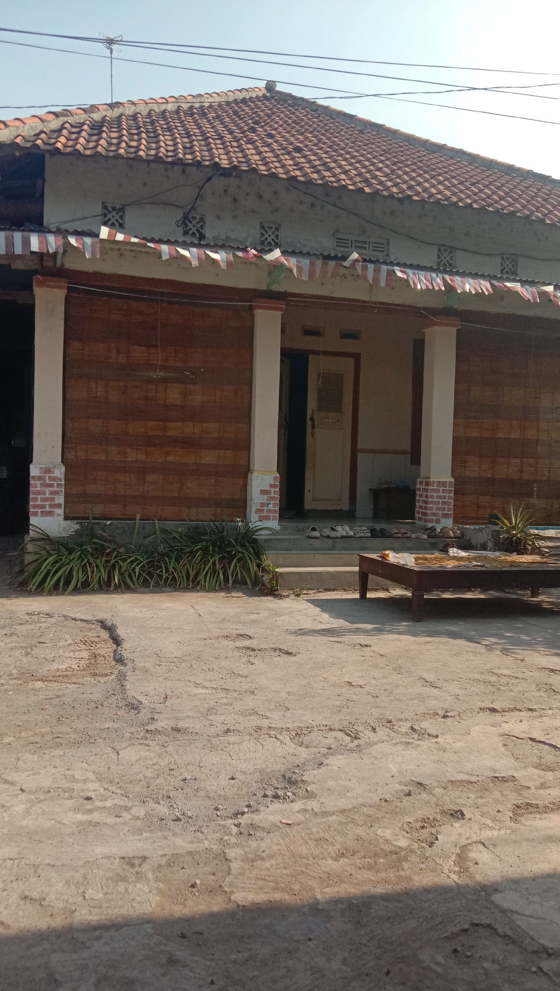The image depicts the exterior of a single-story house in Sidogiri, Indonesia, under a clear sky. The house features a red tile roof and is adorned with a string of small Indonesian flags along the front overhang. The walls are a light color with decorative square vents. The porch is supported by two prominent white columns with brick bases. Bamboo blinds cover a large portion of the front of the house. In front of the house, a row of green plants lines the porch. The ground in front is paved with concrete, showing some cracks and unevenness. To the right of the porch, a low, dark wooden table is placed on the concrete, holding what appears to be dried food items. Several pairs of sandals are neatly arranged on the steps leading to the house entrance. The scene is brightly lit by natural daylight, casting shadows on the ground.