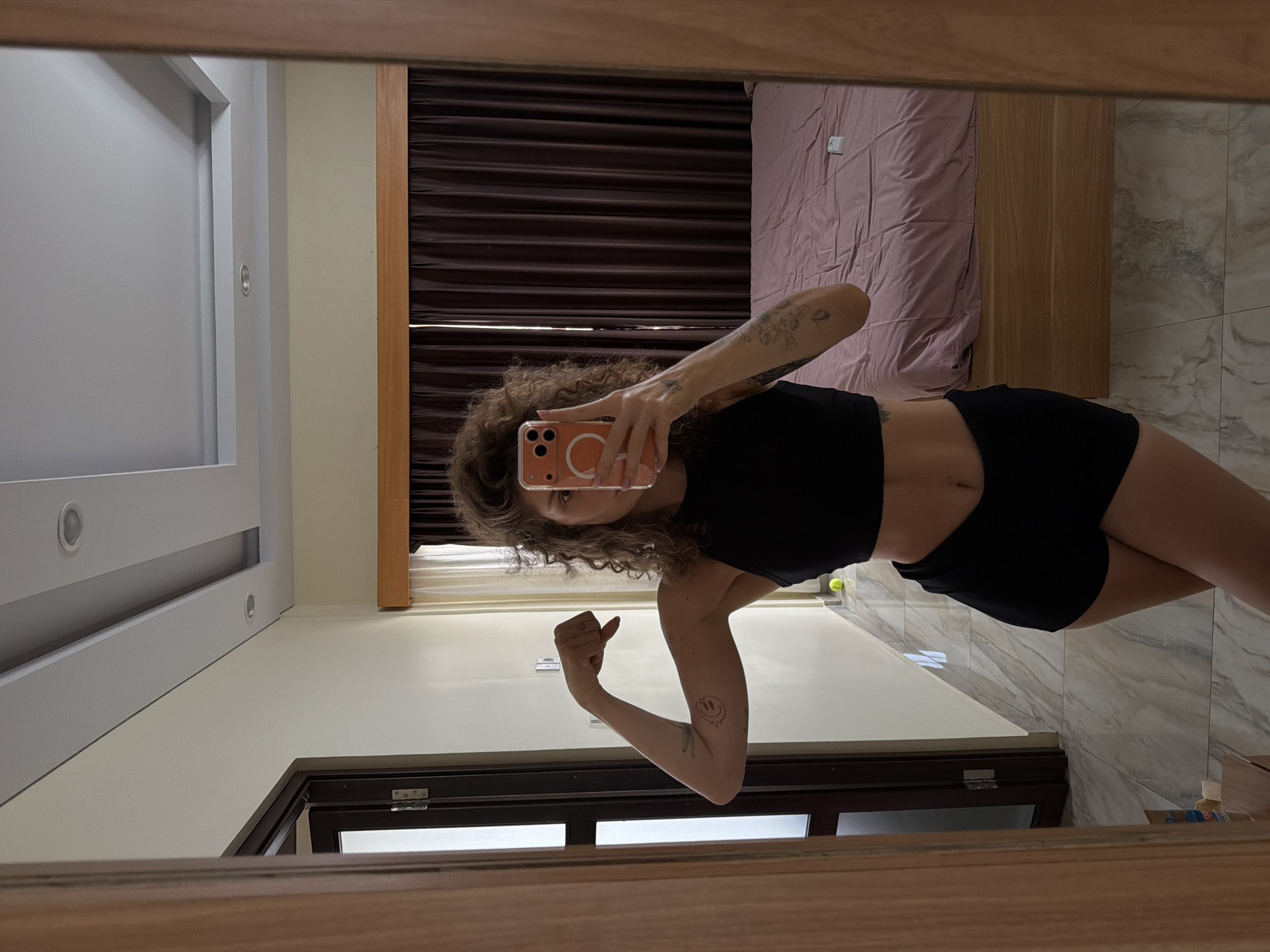 A mirror selfie captures a woman standing in what appears to be a modern bedroom in North Kuta, Indonesia. The woman, with curly brown hair, is wearing a black crop top and black shorts, revealing her midsection. She holds an orange-colored smartphone with a transparent case up to her face, partially obscuring her features, as she takes the photo. Her left arm is flexed, showing off several tattoos, including what appears to be a melting smiley face on her upper arm and floral designs on her forearm.
The background, reflected in the mirror, shows a neatly made bed with light pink sheets and a wooden headboard or frame. Behind the bed are dark brown curtains, partially drawn. To the left of the woman, a white wall is visible with recessed lighting fixtures. The floor is covered with light-colored, greyish-white tiles featuring a marble-like pattern. A dark-framed window is visible in the lower part of the reflection. Other details include a white electrical outlet on the wall above the bed and a small yellow-green ball on the floor in the foreground. The lighting suggests it is daytime.