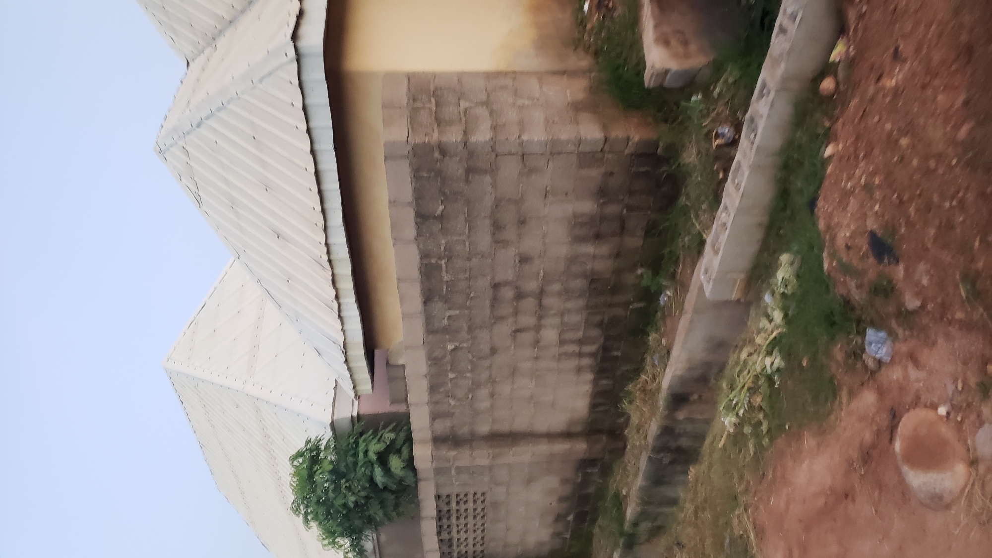 The image depicts a residential area in Jalingo, Nigeria, characterized by a concrete block wall in the foreground, a drainage channel running alongside it, and two buildings with corrugated metal roofs in the background. The wall, constructed from grey concrete blocks, shows signs of weathering and discoloration. To the left of the wall, a section features a decorative pattern of small openings, possibly for ventilation. A mature green tree stands behind the wall, its foliage partially obscuring the building behind it. The buildings themselves have light-colored, sloped roofs with visible rivets. The taller building to the left has a pyramidal roof, while the one to the right has a more elongated, sloped roof. The sky above is a pale, overcast blue. The ground in the foreground is a mixture of reddish-brown soil and sparse green vegetation, with some debris, including plastic bags and dried plant matter, scattered around. A substantial rounded stone or concrete object lies on the soil in the lower left portion of the image. The scene appears to be a street view of houses.