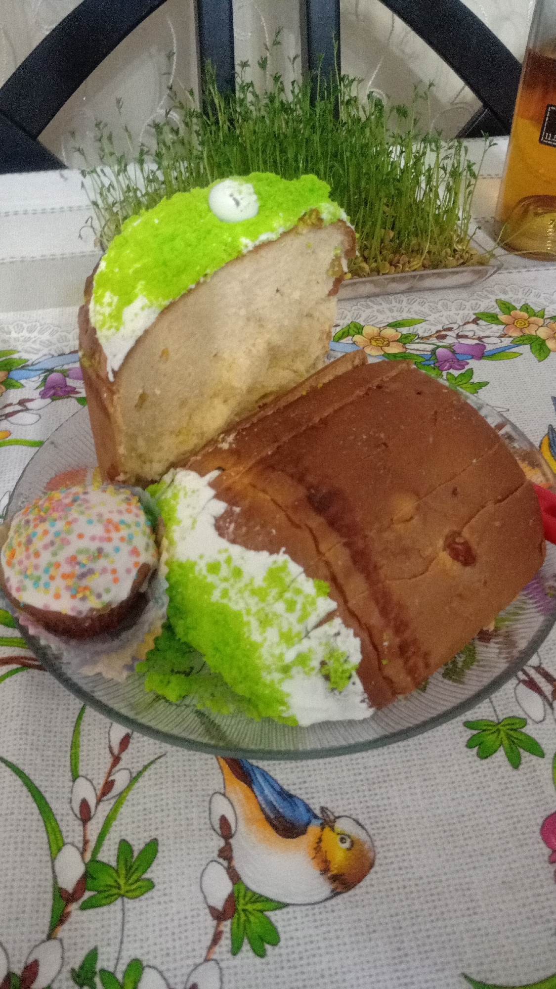 A plate containing a sliced cake with green frosting and a cupcake is placed on a tablecloth with a bird and floral pattern. Behind the plate is a tray of growing sprouts, with a bottle in the background. The location is possibly Yerevan, Armenia, though this is difficult to ascertain from visual information alone. The objects are on a table indoors.