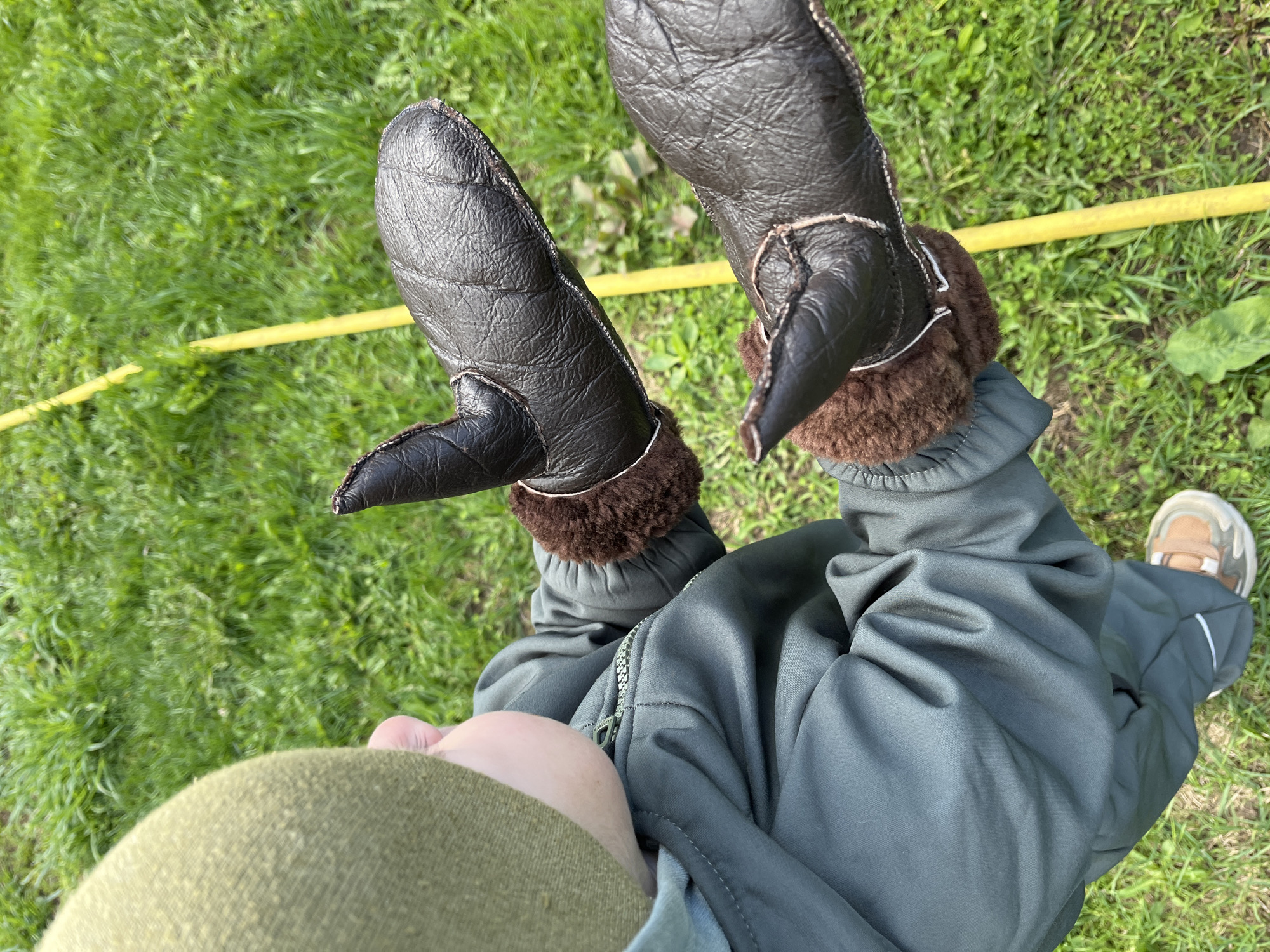 A top-down view shows a child in a dark green winter jumpsuit and unusual dark brown leather mittens with shearling trim, standing on a grassy lawn. A yellow hose lies on the grass near the mittens. The child is also wearing a shoe with brown and white accents. The image was most likely taken in a rural or suburban area with grass. There is no definitive indication of the location being in the city of Chaikovsky, Russia.