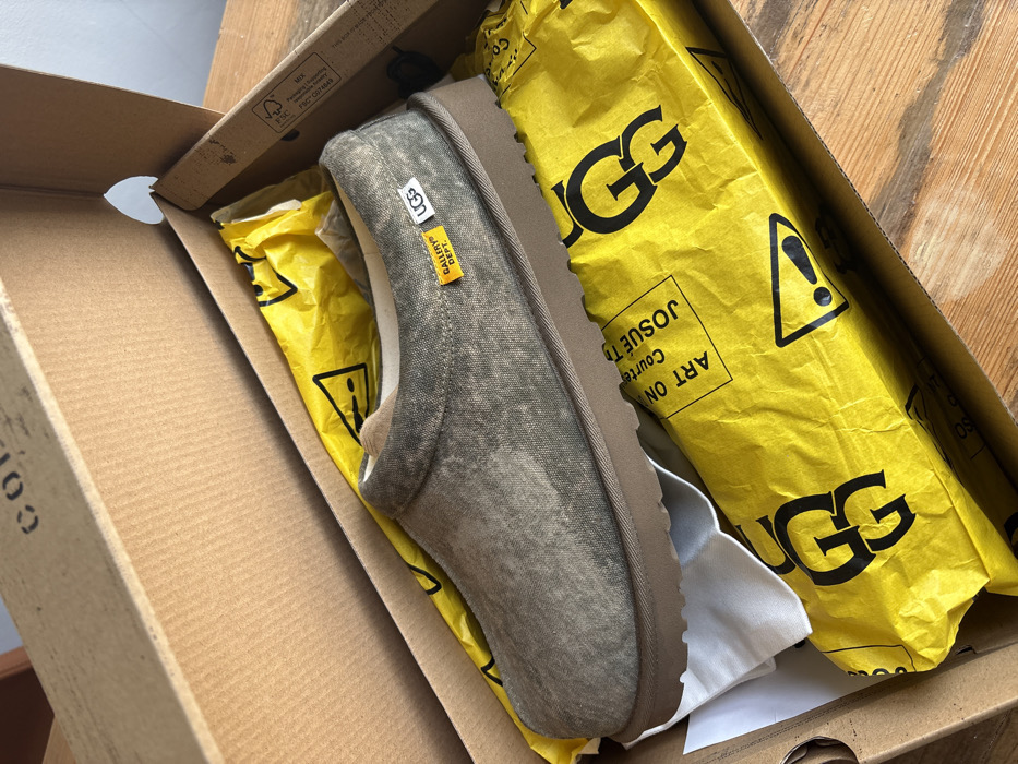The image displays an open, brown corrugated cardboard shoe box resting on a wooden surface in London, United Kingdom. Inside the box, a single, backless slipper is visible, angled towards the right. The slipper's upper is a mottled olive-green or faded grey-green fabric with a distressed appearance, complemented by a light brown, ridged sole. Two small rectangular fabric labels are affixed to the slipper's side: a white label bearing "UGG" in black text and a smaller yellow label marked "GALLERY DEPT." in black. The slipper's interior lining is light-colored and soft. White tissue paper supports the slipper, while crumpled bright yellow tissue paper fills the remaining space in the box. This yellow paper is patterned with black "UGG" logos, exclamation mark warning symbols, and fragmented text, including "ART ON" and "JOSUÉ T". The inner lid of the box shows faint black stencil-like text. A small white label with "FSC MIX" logos and "FSC C074819" is present on the inner right flap of the box. The wooden surface exhibits visible grain, and the scene is brightly lit by ambient light, suggesting an indoor setting. No individuals or active interactions are depicted.