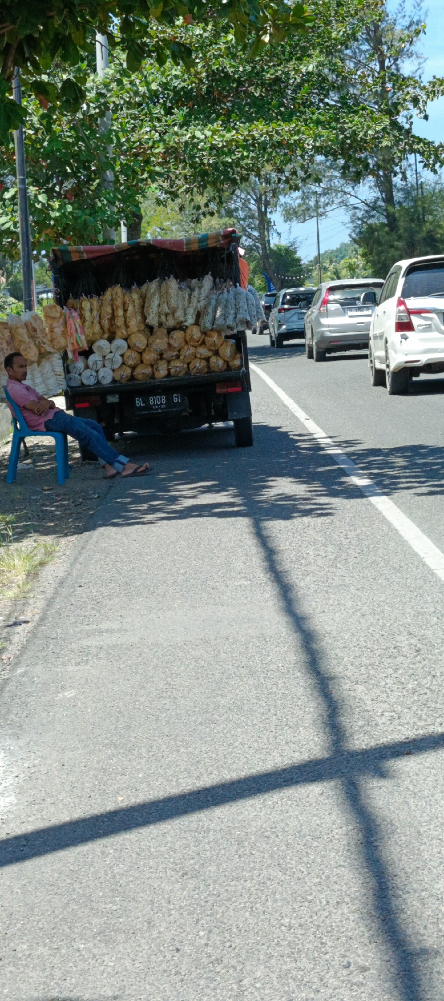 A man sits on a blue plastic chair on the side of a road in Lampisang, Indonesia, a truck filled with snacks parked behind him. The truck, with license plate BL 8108 GI, is draped with a colorful tarp and displays an assortment of dried food products, including what appear to be crackers and chips, hanging and stacked. The man, wearing a pink shirt and blue jeans, appears to be resting or waiting. Several cars are driving on the asphalt road, casting shadows across the scene, which is partially shaded by overhead trees. The environment suggests a sunny day in a semi-rural or coastal area.