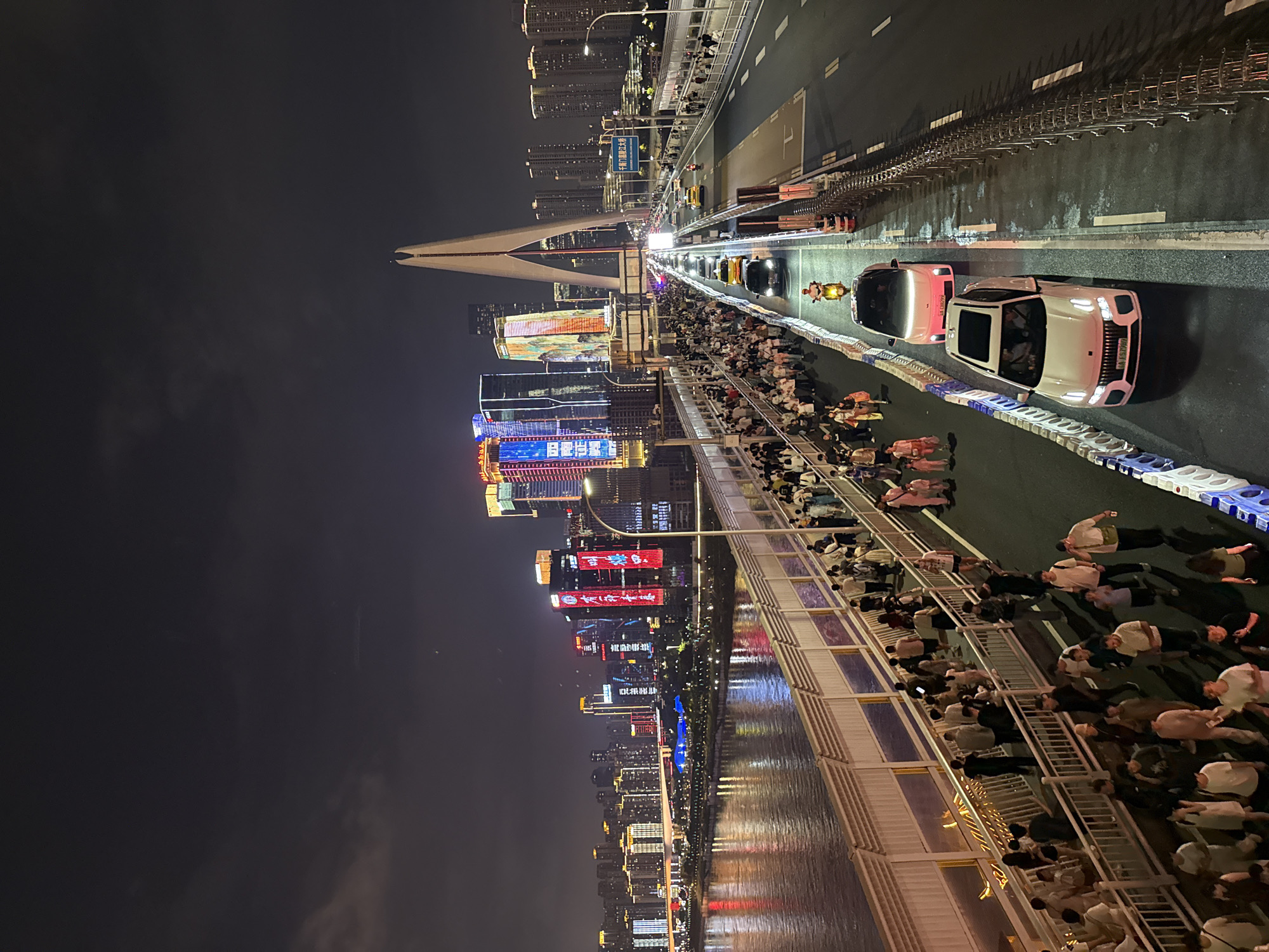 An elevated, wide-angle night shot captures a bustling urban scene in Yuzhong District, China, likely on a pedestrian bridge over a river. The foreground shows a pedestrian walkway densely populated with people strolling. The walkway is separated from a multi-lane road by a white railing. On the road, traffic is moderate, with cars, a scooter, and a motorcycle visible. The road is illuminated by streetlights.
Beyond the road and pedestrian walkway, the city skyline dominates the background. Tall, modern buildings are illuminated with colorful LED lights, creating a vibrant and dynamic spectacle. The river in the foreground reflects the city lights, adding to the visual richness of the scene. A distinctive modern bridge with a prominent arch structure spans the river, connecting different parts of the city. The sky is dark and cloudy, indicative of nighttime.
The overall impression is one of a lively, modern metropolis at night, with significant pedestrian and vehicular activity. The presence of numerous people suggests a popular attraction or event.