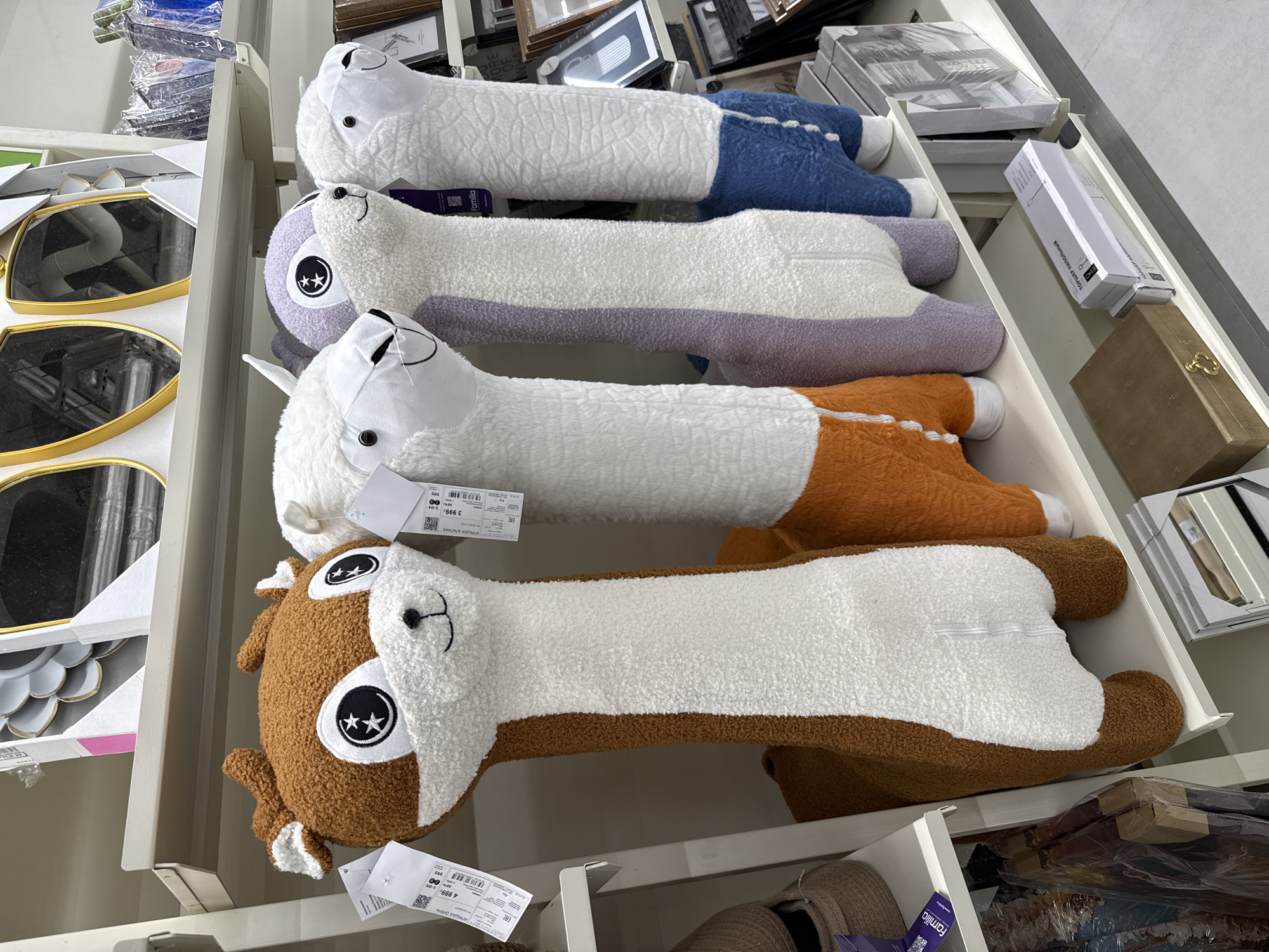The image depicts a retail shelf stocked with elongated, plush animal toys, possibly in a store located in Izhevsk, Russia, based on price tags in Russian. From top to bottom, the toys are: a white llama with blue "legs", a white llama with purple "legs", a white llama with orange "legs", and a brown reindeer-like creature with white areas. Each toy has attached price tags. Other retail merchandise, including mirrors and framed pictures, are visible on adjacent shelves. The environment appears to be a well-lit indoor setting within a retail establishment.