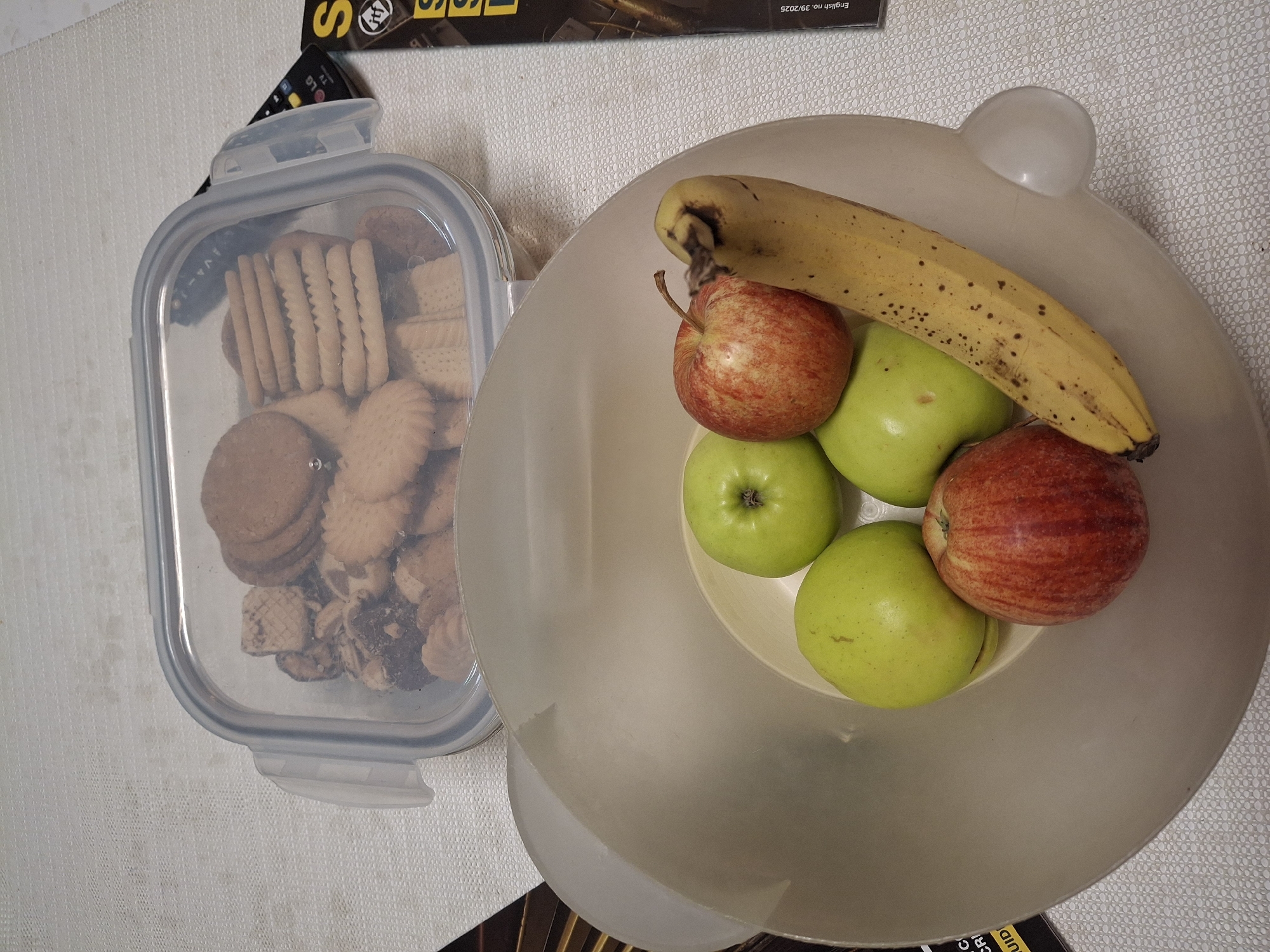 The image shows a container of cookies and a bowl of fruit. The bowl contains a banana and four apples. The cookies are a variety of shapes and sizes. The scene is indoors. The location is not specified and cannot be identified as being in Eemshaven, Netherlands. There are no people visible in the image. There are no actions or interactions depicted. The environment is a white, patterned surface, possibly a tablecloth or countertop.