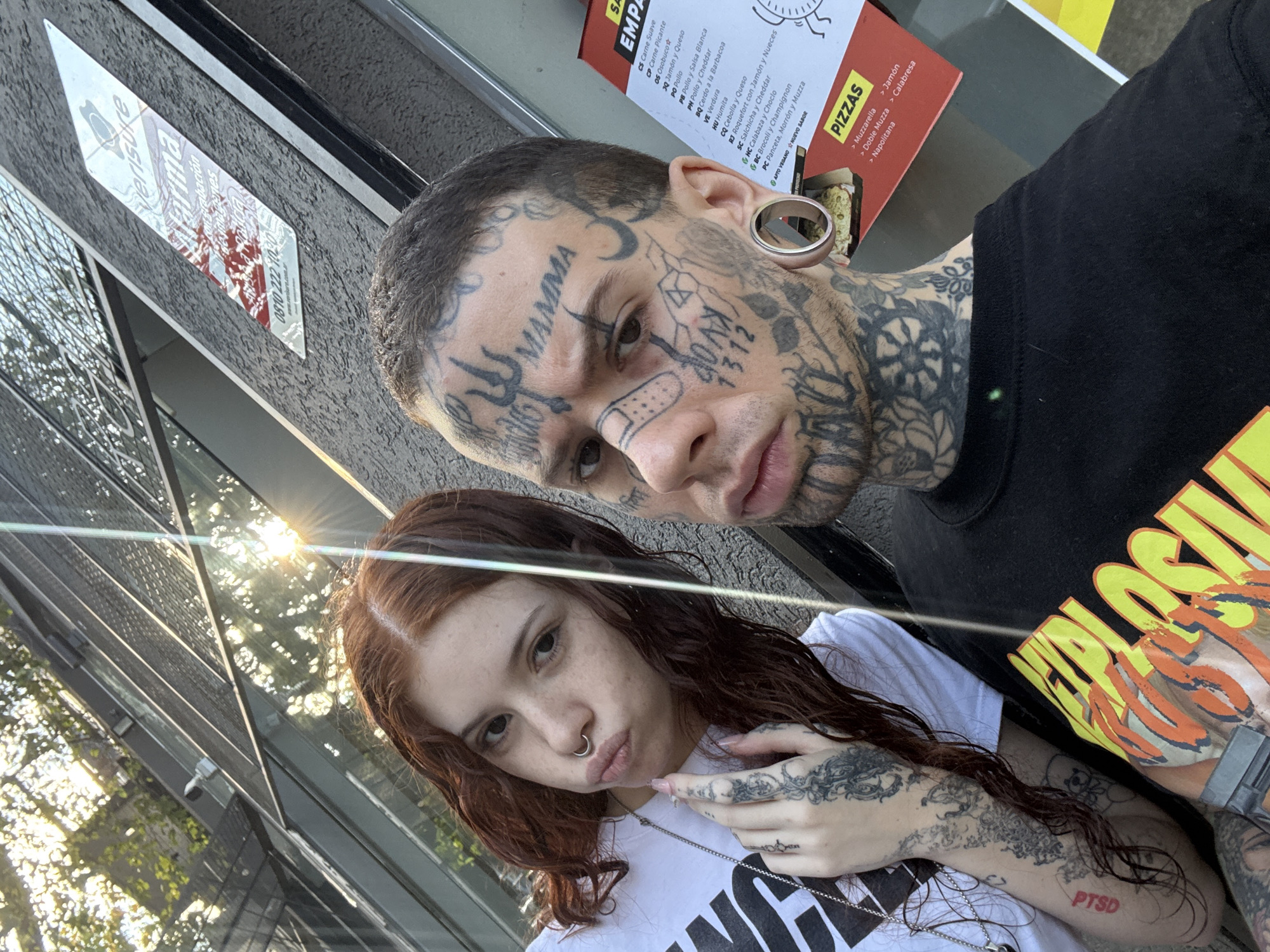 The image shows two people, one male and one female, posing for a selfie. The male has numerous face tattoos, a bandage on his nose, and a stretched earlobe. He wears a black t-shirt with yellow and orange graphics. The female has red hair, a nose ring, and arm tattoos. She wears a white t-shirt.
The background includes the exterior of a building. A sign on the building indicates a security system by Verisure, with the phone number 0810 222 1020. The sign also has the word "Verisure" and "congrabación de imagenes" written on it. A menu for "Empa" is visible, listing various fillings, including "Carne Suave," "Carne Picante," "Jamón y Queso," "Pollo," and "Verdura." It also lists pizza options such as "Muzzarella" and "Jamón."
The location is suggested to be in the city of Buenos Aires, Argentina, given the presence of Argentinian brands.