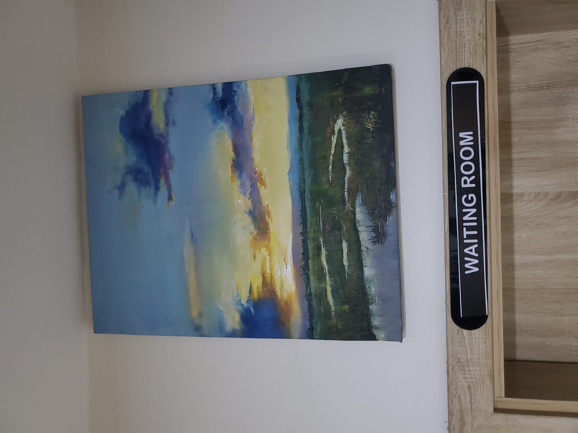 A painting of a sunset over a marshy landscape hangs on a white wall. The painting depicts a sky with vibrant hues of orange, yellow, and purple clouds, transitioning to a blue sky above. A dark, green marsh with reflective water channels stretches across the lower half of the painting, with distant blue hills forming the horizon. A signature and date are visible in the bottom right corner of the painting. Below the painting, a black sign with white lettering reads "WAITING ROOM" and is mounted on a light-colored wooden door frame. The image captures a scene within the city of Jos, Nigeria, in a waiting area.