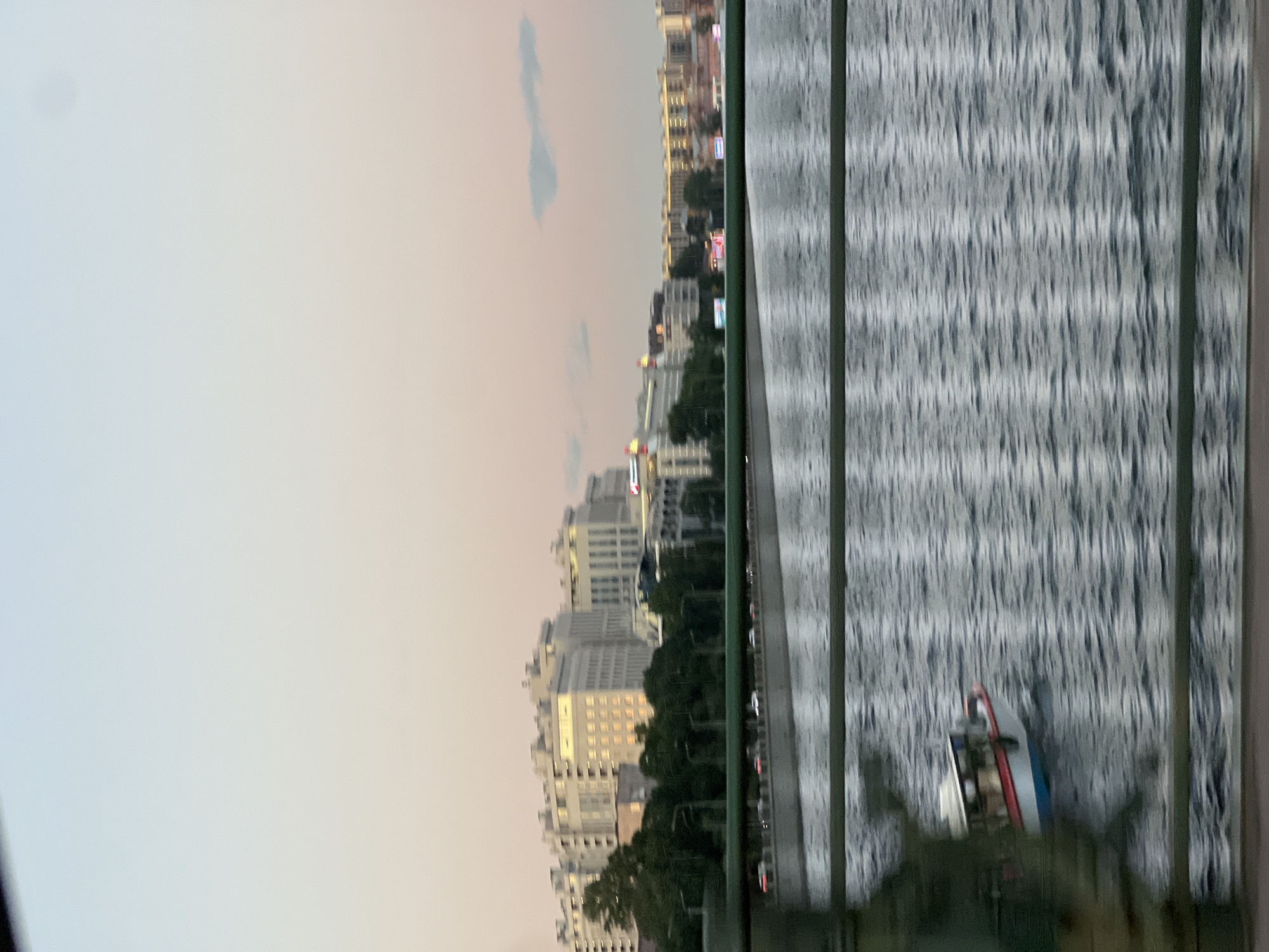 This is a landscape photograph taken from a moving vehicle, likely a train or car, looking across a body of water towards a city skyline. The image is blurred due to motion, creating streaks of light and indistinct shapes. In the foreground, a portion of a dark green railing is visible, along with a faint suggestion of a person or object on the left. The water in the middle ground is choppy, with ripples and reflections. A small boat with a red and white hull is visible on the water, moving from left to right. In the background, a city skyline is depicted with several multi-story buildings, appearing to be residential or office complexes. Trees line the waterfront between the water and the buildings. The sky is a soft gradient of pale blue and pink, suggesting either dawn or dusk. The overall impression is of a city viewed from its river or canal. Given the architecture and the presence of the canals, the location is likely Saint Petersburg, Russia.