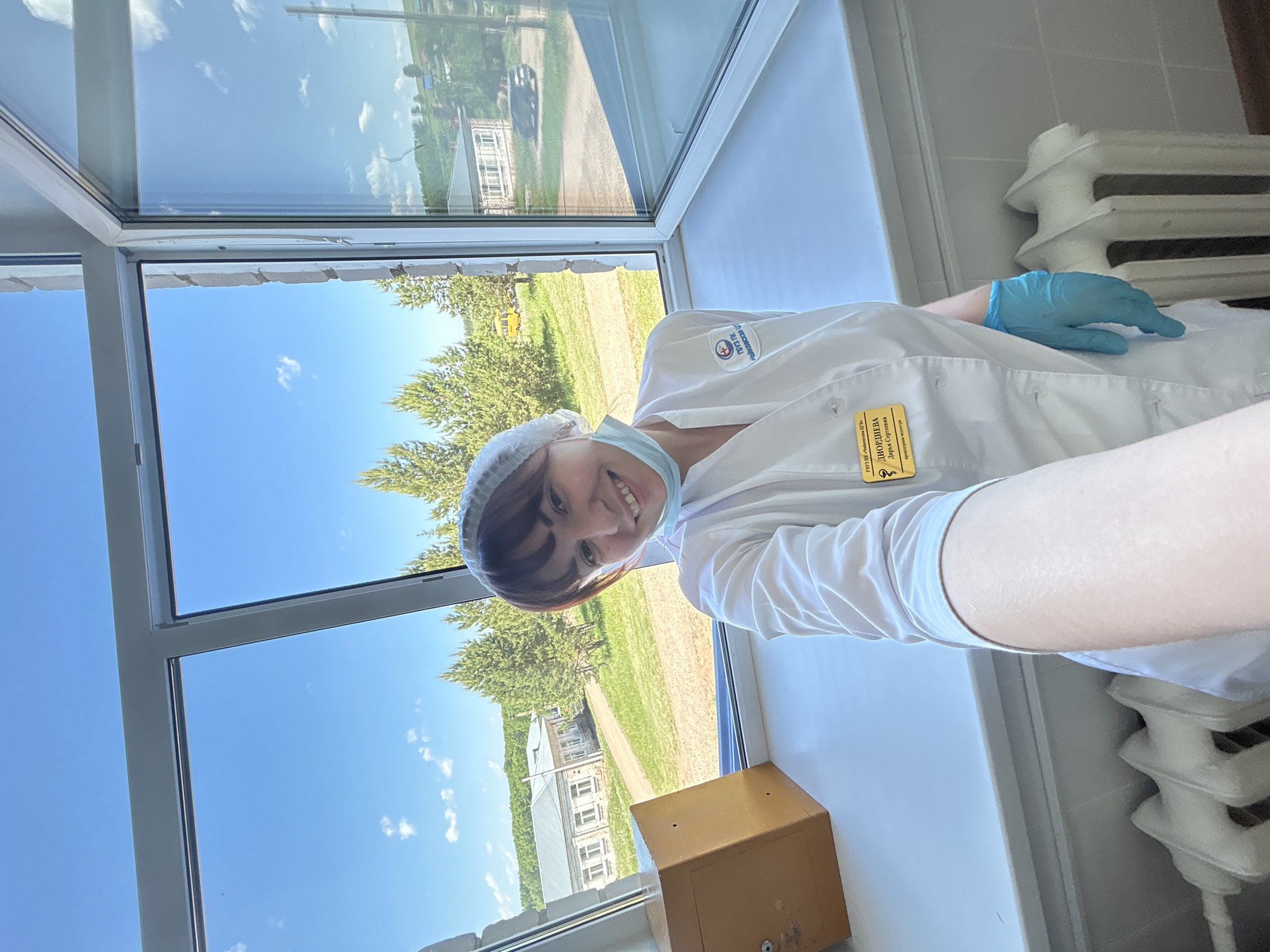 A young woman, likely a healthcare worker, poses for a selfie indoors next to a window. She wears a white medical coat, a surgical cap, and a blue surgical glove on her right hand. Her identification badge is visible on her coat. A radiator is visible to her right. Through the window, the sky is blue, some trees are visible, and what appears to be a hospital building or similar institutional building is visible, and a brown box is also visible. According to the OCR, her last name is Diordieva. The text also identifies the location as the city of Choyka, Russia.