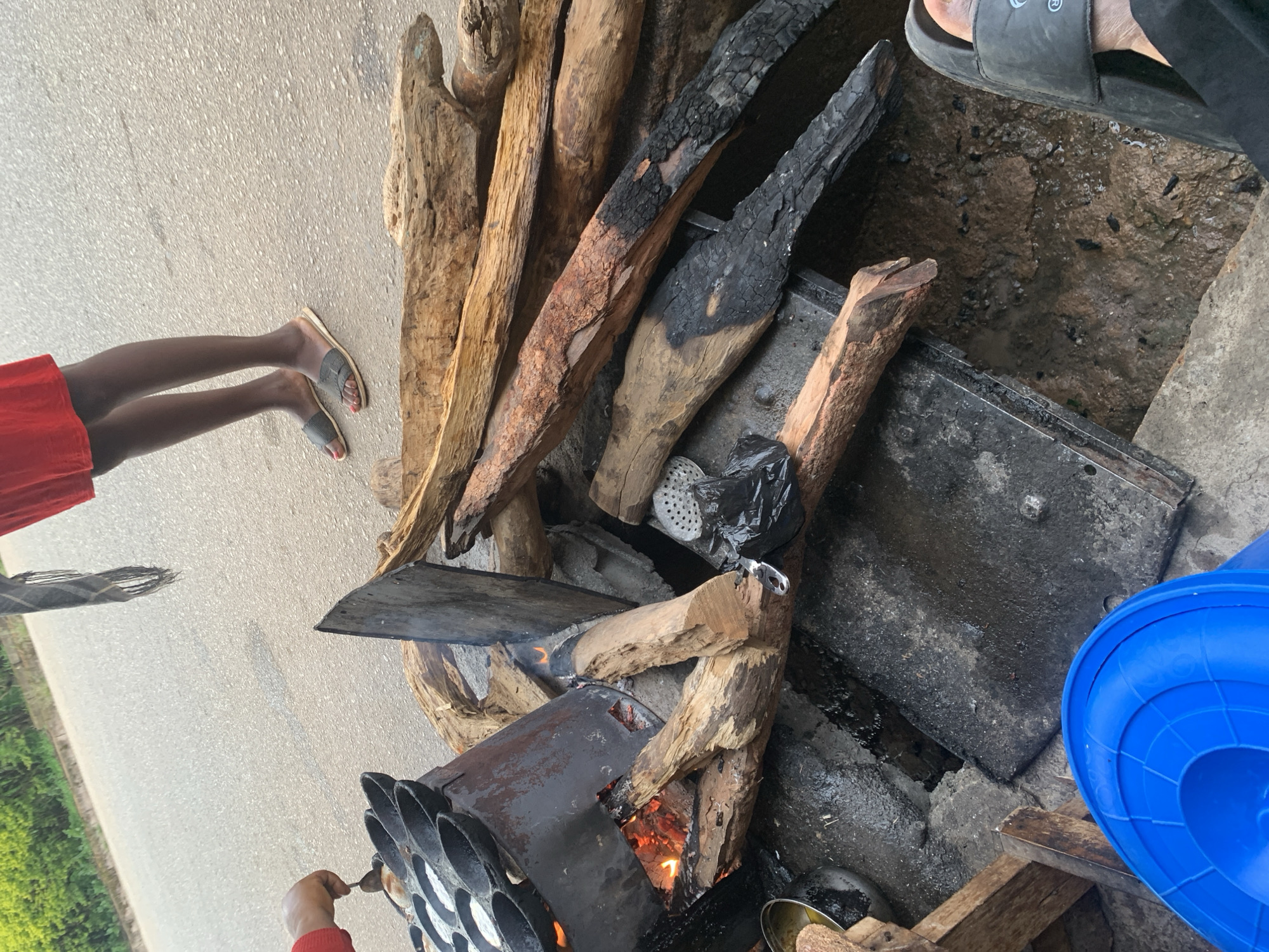 This image depicts an outdoor cooking scene likely in Jos, Nigeria. In the foreground, a person's hand in a red sleeve is using a spoon to prepare food in a cast-iron pan with multiple indentations, which is placed over a fire. Several pieces of firewood, some charred, are stacked and used as fuel for the fire. Adjacent to the cooking area, a metal colander sits in a black plastic bag. The ground appears to be damp and uneven, possibly a dirt or gravel surface. In the upper portion of the image, another person's legs are visible, wearing a red garment and dark sandals, standing on a paved surface that could be a roadside. A blue plastic container is also present in the lower left corner. The overall impression is a rustic, possibly informal, food preparation setup in an urban or peri-urban environment.