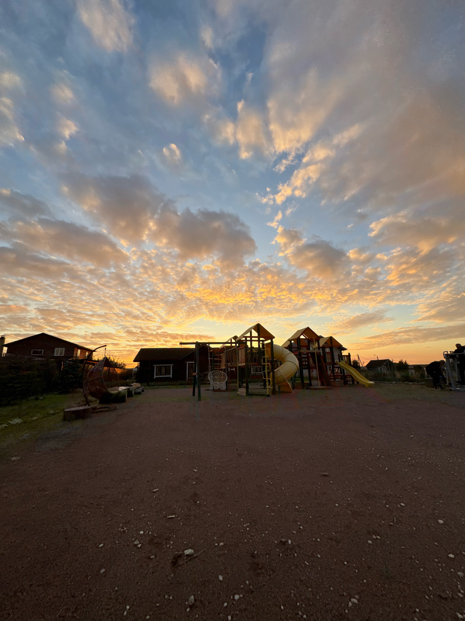 This image depicts an outdoor playground area in Kempelevo, Russia, during a vibrant sunset. The sky is filled with dramatic, wispy clouds illuminated in shades of orange, yellow, and blue. In the foreground, a dirt ground surface is visible with scattered small stones and debris. To the left, a wooden house with dark siding and white trim stands, partially obscured by trees and bushes. Adjacent to the house, a unique, circular, woven hanging chair is suspended from a metal frame, resting on a tree stump. Further to the right, another dark-sided wooden building with white trim is present.
Dominating the center of the image is a colorful playground structure featuring yellow slides, including a curved tube slide and a straight slide, along with climbing elements and platforms. In the far right of the frame, two individuals are visible using outdoor exercise equipment, with one person bent over. The overall scene suggests a residential or communal outdoor space at the end of the day.