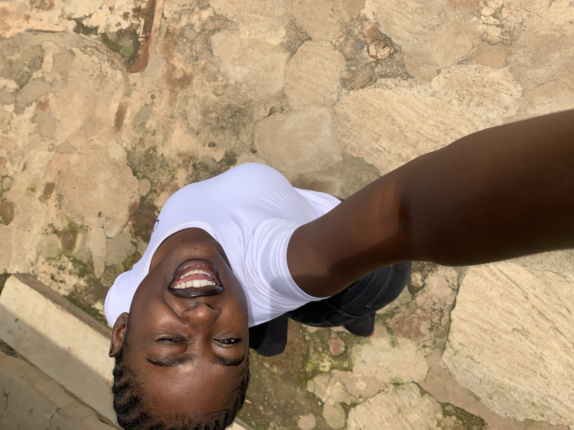 In Rafin Par, Nigeria, a close-up, high-angle selfie shows a young Black woman with braided hair smiling widely. She is wearing a white, short-sleeved shirt and black pants. Her left eye is closed, and her teeth are visible. She is holding the camera, suggesting she is taking the picture herself. The background is an outdoor environment with textured ground made of rough, irregular stones, some of which appear weathered and stained with green moss or algae, indicating an aged or natural setting. Some stone structures, possibly part of a building or pathway, are visible in the upper left portion of the image. The lighting suggests a daytime scene.