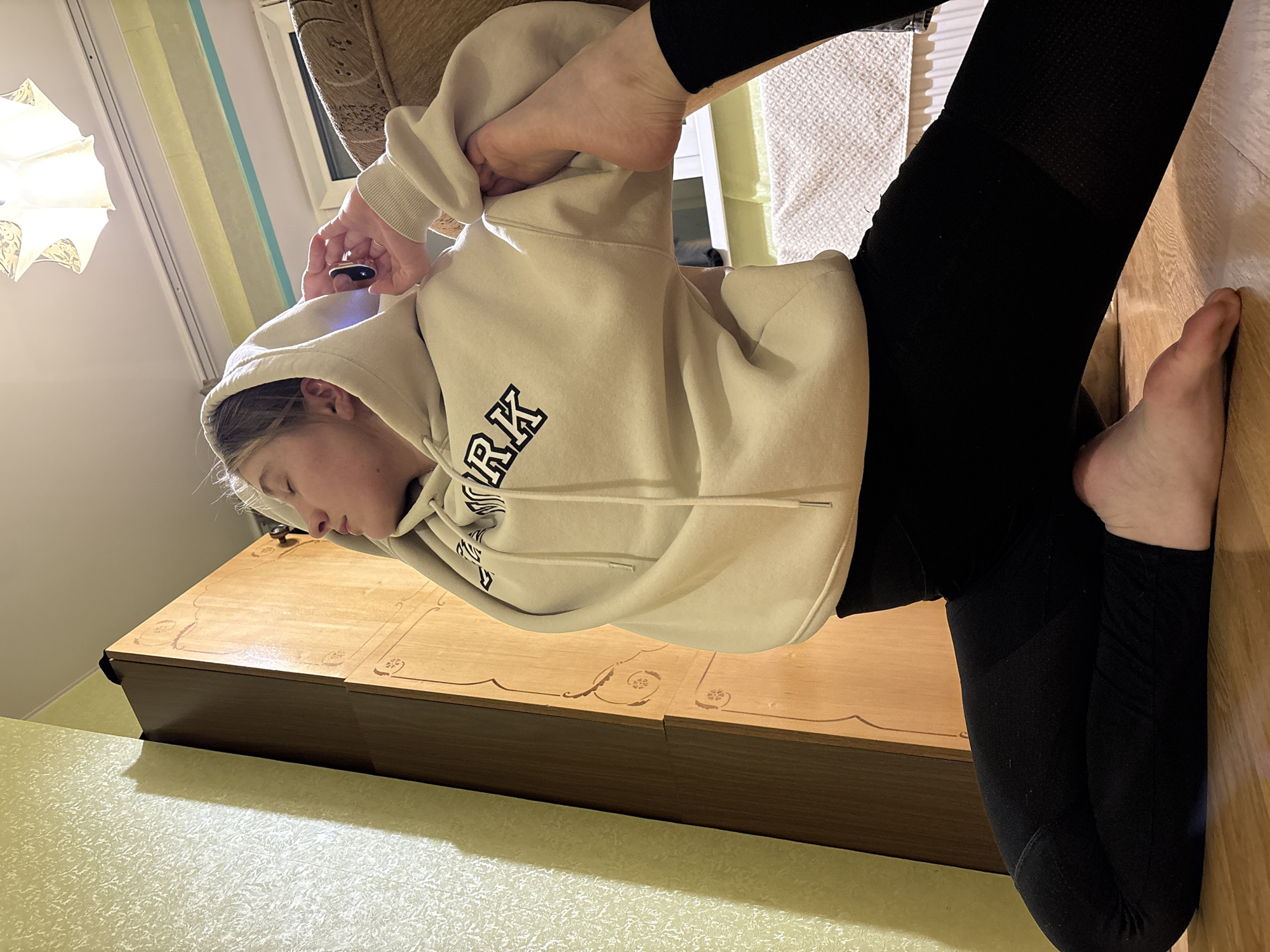 A young person in a beige hoodie and black leggings is seated on a light-colored wooden dresser, with one bare foot on the wooden floor. The location is indoors, and the presence of curtains and a light fixture suggests a residential setting. The person appears to be using a small black object near their face, possibly a mobile phone. The scene is well-lit, and the environment is domestic. The image does not provide any indication of the location being Izhevsk, Russia.