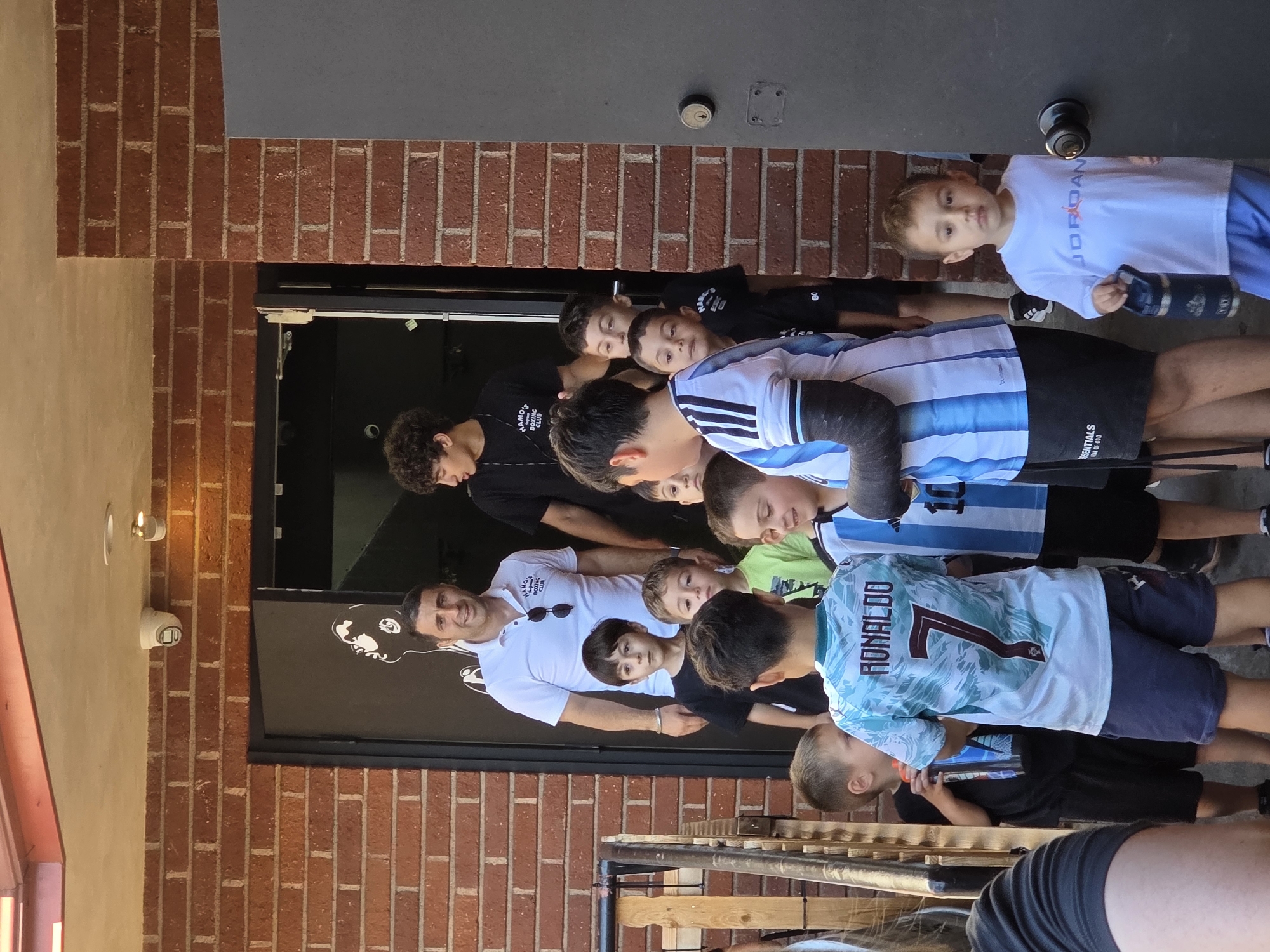 A group of boys, most wearing soccer jerseys, stand outside a brick building with a dark entrance. A man in a white polo shirt stands behind some of the boys, looking towards the camera. The building has a dark door with a mural of a face on it. There are two security cameras mounted on the brick wall above the entrance. One of the boys in the front is wearing a light blue and white jersey with "RONALDO 7" on the back. Another boy to the right is wearing a white t-shirt with "JORDAN" logo. The scene appears to be outdoors on a sunny day. The location is the city of Glendale, United States of America.